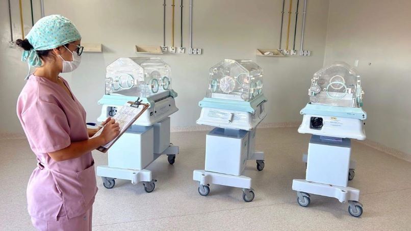 Profissional de saúde usando uniforme rosa, touca e máscara observa e anota em uma prancheta diante de três incubadoras neonatais em uma sala hospitalar clara e equipada.