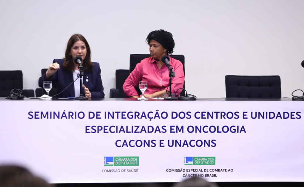 Duas mulheres estão sentadas em uma mesa de seminário. À esquerda, uma delas fala ao microfone, enquanto a outra escuta com atenção. À frente da mesa, há uma faixa com os dizeres “Seminário de Integração dos Centros e Unidades Especializadas em Oncologia – CACONs e UNACONs”, com logotipos da Câmara dos Deputados e das comissões de saúde e combate ao câncer no Brasil.