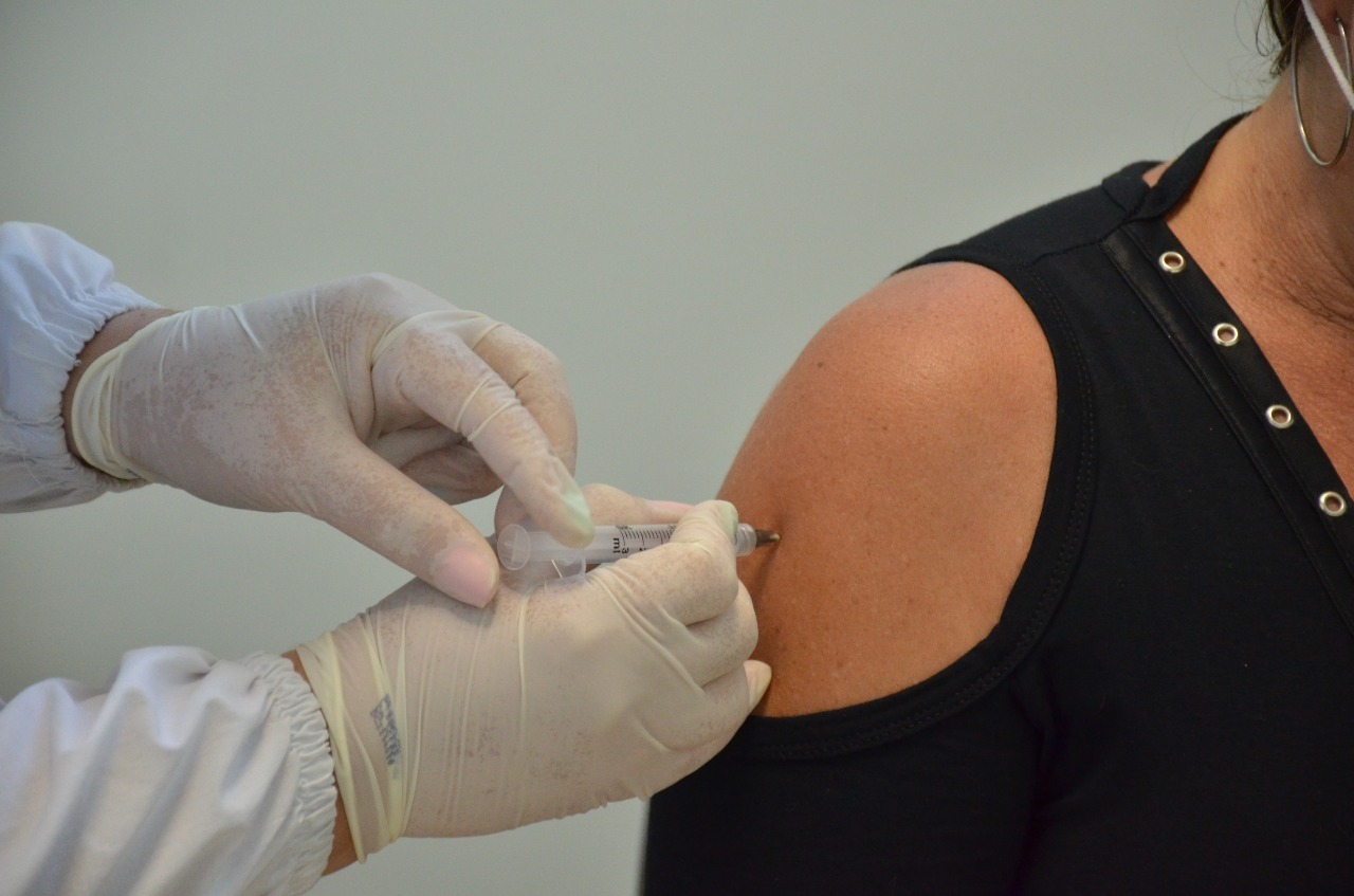Profissional da saúde com luvas aplicando uma vacina no braço de uma mulher vestindo blusa preta. A imagem retrata o momento da imunização com seringa inserida no braço da paciente.