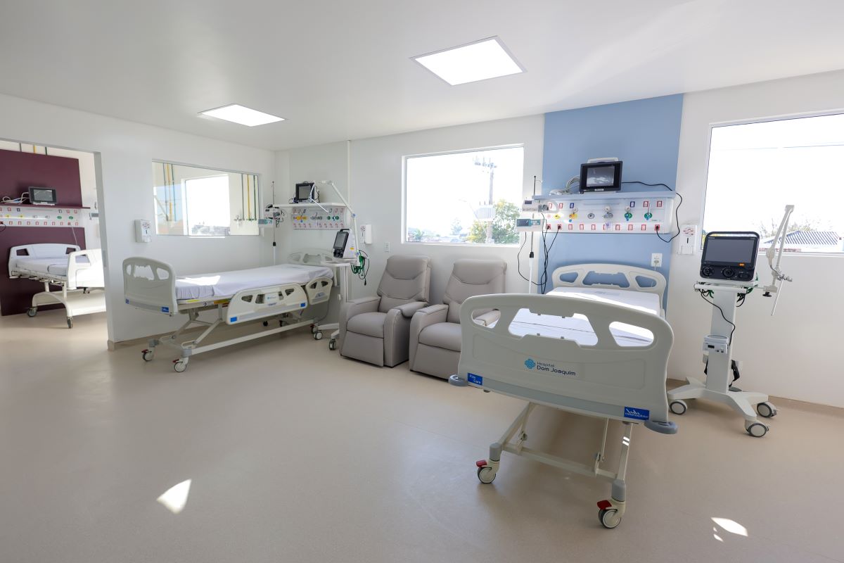 A imagem mostra uma ala de UTI pediátrica moderna e bem iluminada.  O ambiente conta com:  Três leitos hospitalares com grades de proteção e rodízios.  Poltronas reclináveis ao centro, provavelmente para acompanhantes.  Equipamentos médicos como monitores cardíacos, respiradores e painéis de gases medicinais e tomadas.  Janelas amplas que permitem entrada de luz natural, deixando o espaço mais acolhedor.  Piso claro e paredes em tons neutros, transmitindo sensação de limpeza e organização.  O espaço parece recém-inaugurado e preparado para atendimento intensivo pediátrico.