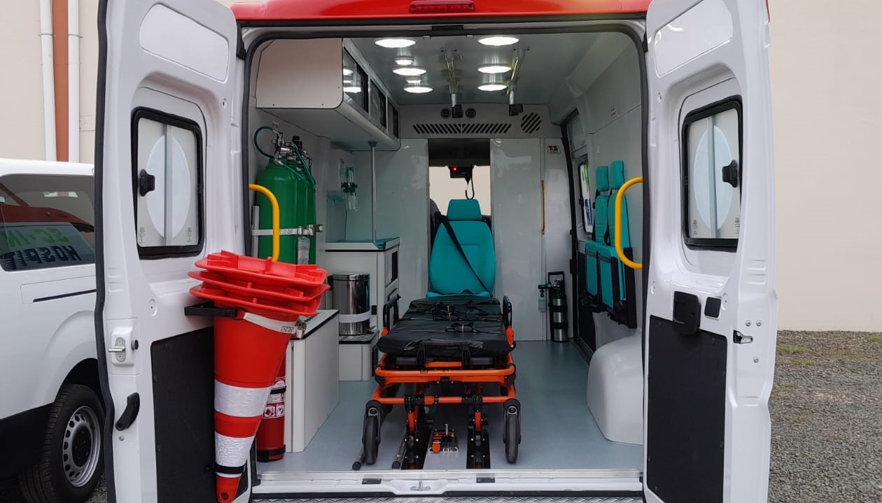 Vista interna de uma ambulância equipada com maca central, cadeiras laterais, cilindros de oxigênio, armários com suprimentos médicos e iluminação no teto. Cone de sinalização e extintor visíveis na porta traseira aberta.