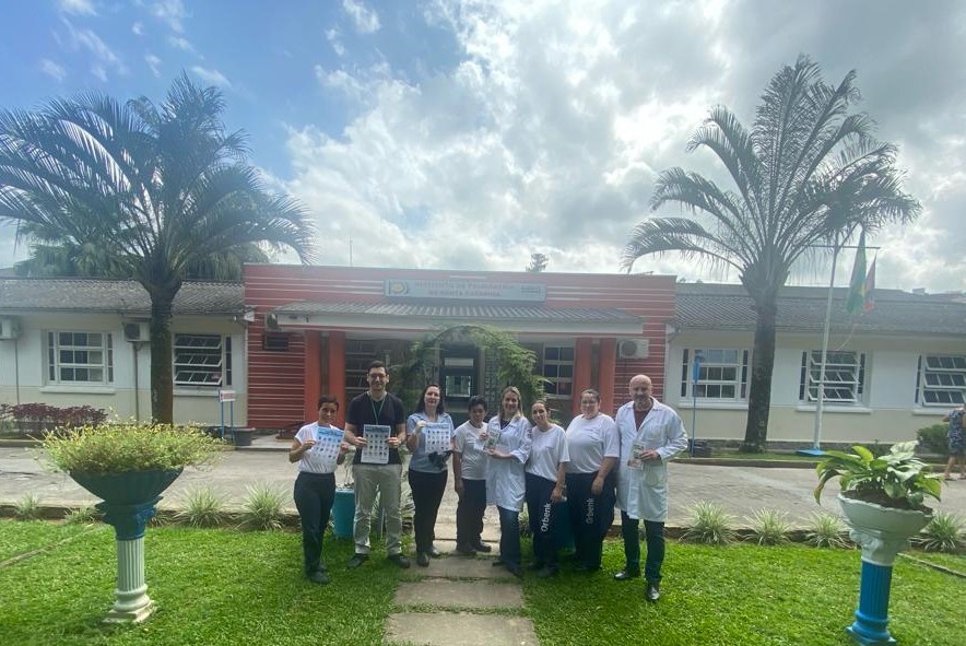 Grupo de oito pessoas posa para foto em frente a um prédio com fachada branca e laranja, identificado como Instituto de Psiquiatria. Eles seguram panfletos ou cartelas, alguns vestindo jalecos brancos e outros roupas casuais. Há coqueiros e vasos de plantas ornamentando o gramado em primeiro plano, sob um céu parcialmente nublado.