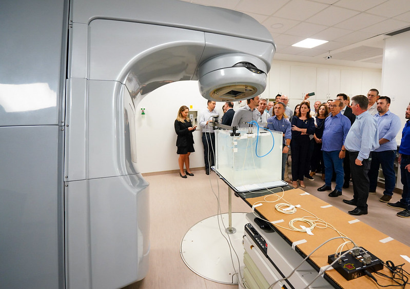 Fotografia colorida de uma sala moderna com equipamento de radioterapia em destaque à esquerda. O aparelho é grande e metálico, voltado para uma bancada com tanque de água e sensores. À direita da imagem, um grupo de pessoas — incluindo profissionais da saúde, autoridades e visitantes — observa a apresentação. O ambiente é bem iluminado, com piso claro e teto com iluminação embutida.