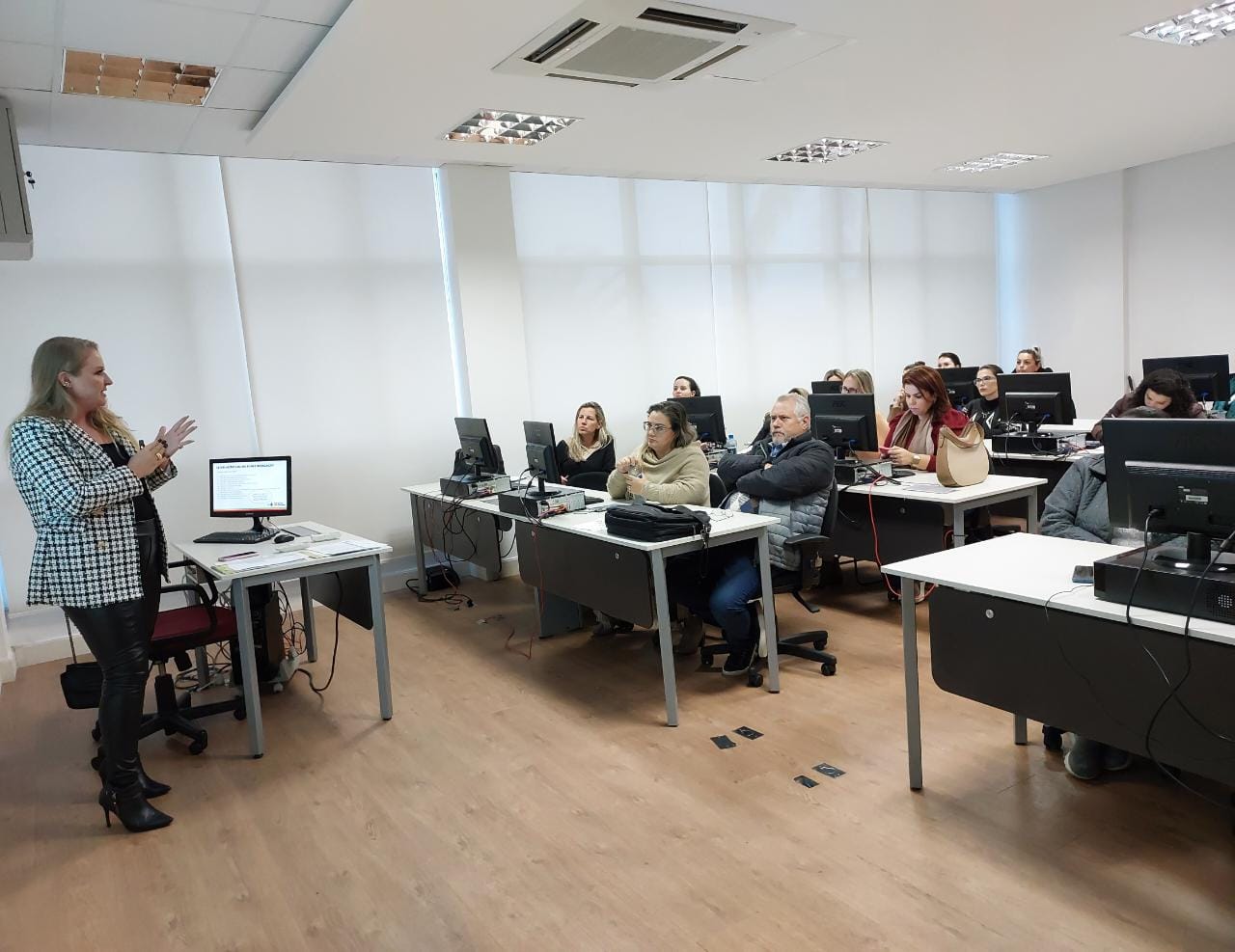 Mulher de cabelo loiro apresenta conteúdo em pé ao lado de um computador, enquanto cerca de 12 participantes, sentados em mesas com computadores, acompanham a capacitação em uma sala moderna e bem iluminada.