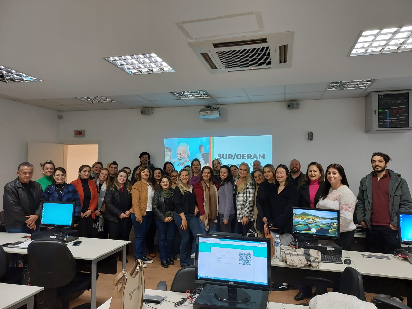 Cerca de 30 pessoas, entre homens e mulheres, estão em pé, lado a lado, sorrindo para a foto em uma sala com computadores. Ao fundo, é projetado um slide com o título 'SUR/GERAM' e a imagem de um idoso sorrindo. O ambiente é de uma capacitação ou treinamento, com cadeiras, mesas, computadores ligados e atmosfera profissional.