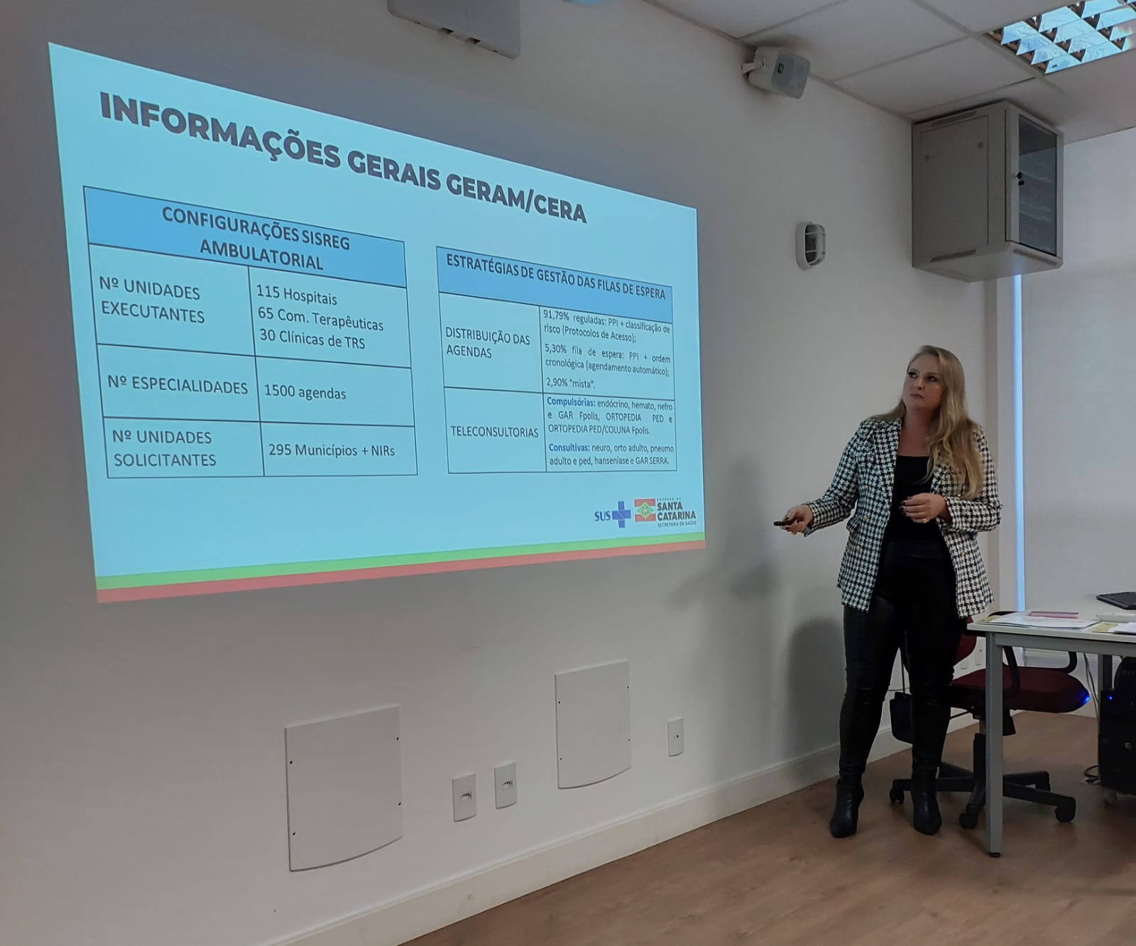 Mulher em pé ao lado de uma mesa, segurando controle remoto, apresenta slide com o título 'Informações Gerais GERAM/CERA', mostrando dados como 115 hospitais executores, 1500 agendas, 295 municípios solicitantes e estratégias para gestão das filas de espera no SUS, durante capacitação em sala de aula.