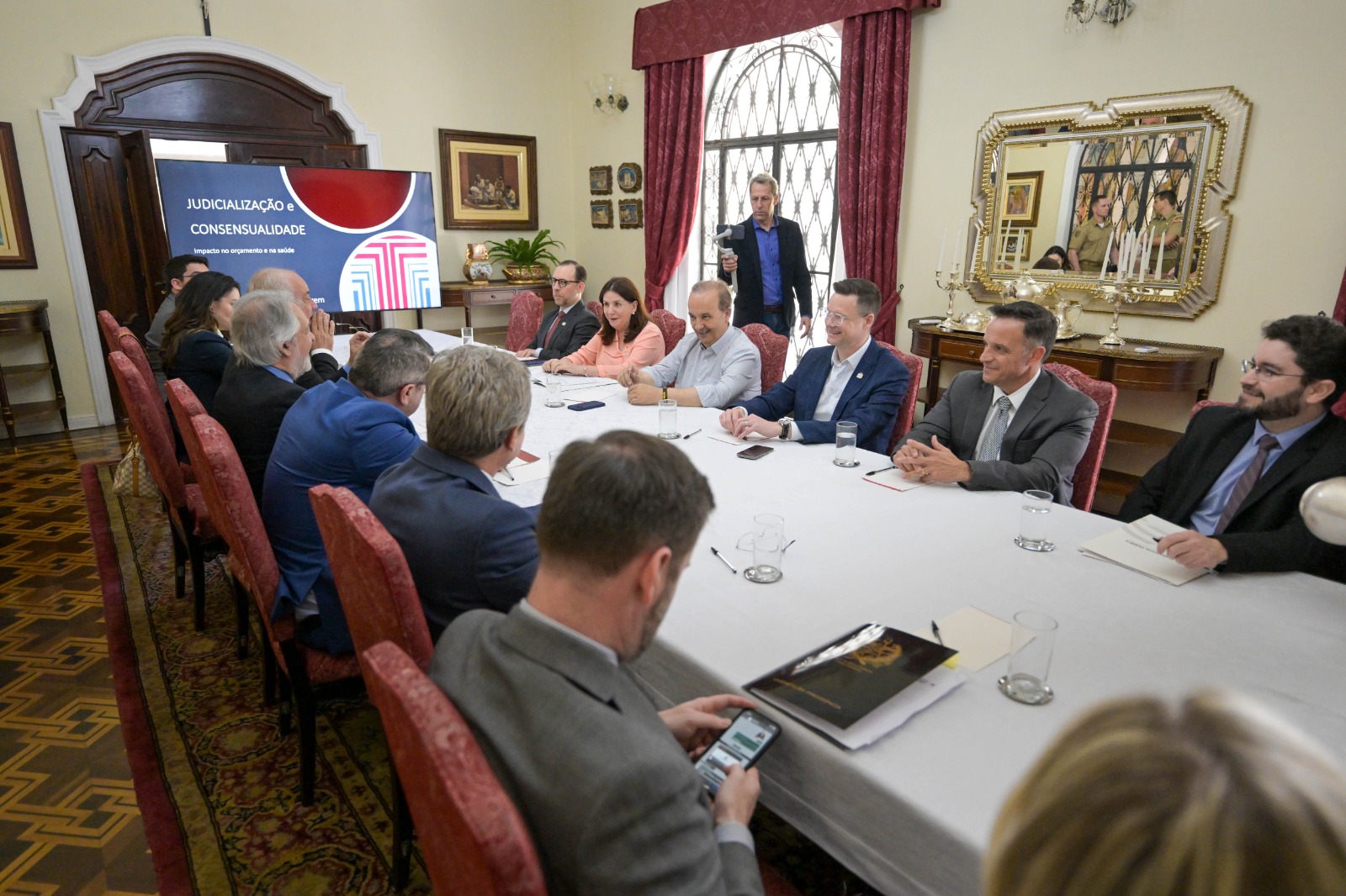 Reunião formal em uma sala com decoração clássica, onde autoridades e representantes de diferentes setores participam de uma mesa oval. Ao fundo, uma tela exibe o tema “Judicialização e Consensualidade: impactos na governança e na saúde”.
