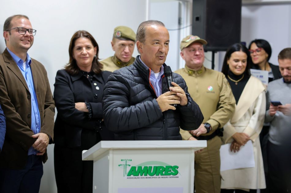 Homem discursa ao microfone durante evento oficial na sede da AMURES, em frente a um púlpito. Atrás dele, estão outras autoridades civis e militares, incluindo policiais, políticos e representantes da comunidade. A cena ocorre em ambiente interno, com clima formal e institucional.