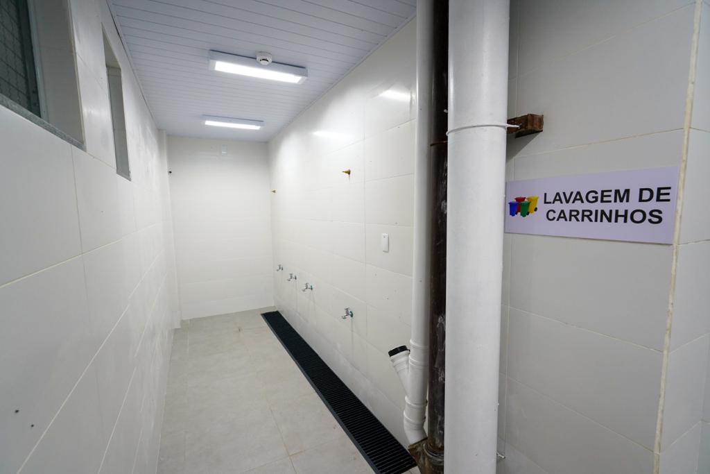 Corredor interno revestido com azulejos brancos, contendo várias torneiras fixadas na parede para lavagem de carrinhos. À direita, há uma placa com a inscrição "Lavagem de Carrinhos" e ícones de coletores coloridos. O ambiente é bem iluminado com luminárias no teto e possui sistema de escoamento no chão.