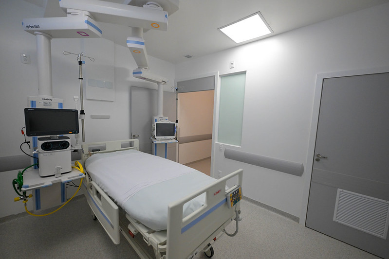 Leito hospitalar equipado com monitor multiparamétrico, suporte de soro, respirador e equipamentos de última geração em uma sala branca, limpa e bem iluminada. O ambiente é moderno, com acabamento em tons claros e portas cinzas.