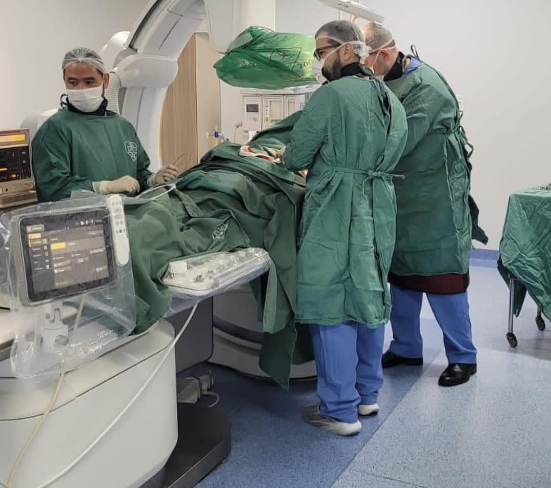 A imagem mostra uma equipe médica realizando um procedimento em um paciente dentro de uma sala de cirurgia ou hemodinâmica.  Três profissionais de saúde estão vestidos com aventais estéreis verdes, máscaras e toucas, posicionados ao redor do paciente, que está deitado em uma maca sob um equipamento médico avançado. Há monitores de sinais vitais à esquerda e outros equipamentos estéreis cobertos por plásticos protetores. A cena transmite um ambiente hospitalar de alta complexidade, possivelmente um procedimento de implante de marcapasso ou outro tipo de intervenção cardiovascular.