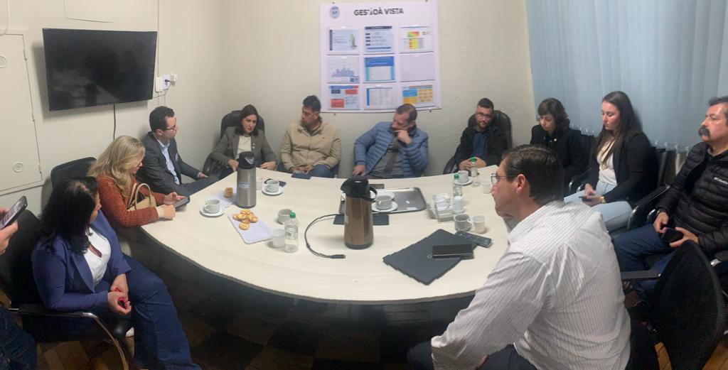 Reunião com cerca de 13 pessoas sentadas em torno de uma mesa oval em uma sala de reuniões pequena. Na mesa, há garrafas térmicas, copos, papéis, biscoitos e garrafas d’água. Na parede ao fundo, cartazes com gráficos e indicadores estão fixados sob o título ‘Gestão à Vista’. As pessoas estão atentas, algumas fazendo anotações ou segurando celulares, em clima de concentração e diálogo.
