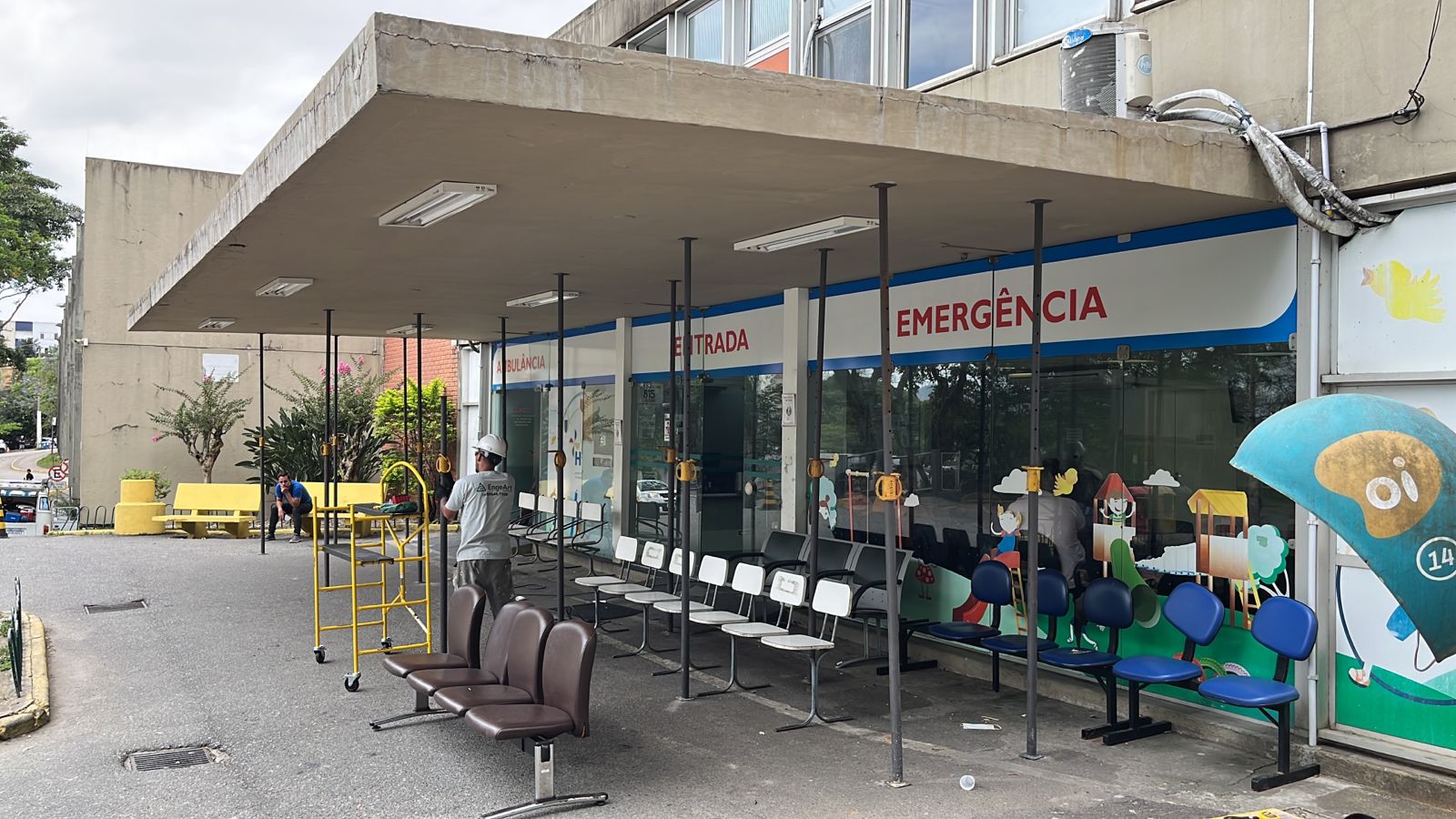 A imagem mostra a entrada de um hospital, identificada pelas palavras “ENTRADA” e “EMERGÊNCIA” acima de portas de vidro, com letreiros também indicando acesso para ambulâncias. A marquise de concreto sobre a entrada está sustentada por vários escoramentos metálicos verticais, sugerindo manutenção ou reparo estrutural. Na área externa, há fileiras de cadeiras de espera, algumas azuis e outras marrons, além de um trabalhador usando capacete e uniforme cinza próximo a um andaime amarelo. Ao fundo, é possível ver bancos amarelos, árvores e parte do estacionamento. À direita, um antigo telefone público no formato de “orelhão” azul e dourado está instalado próximo à parede.