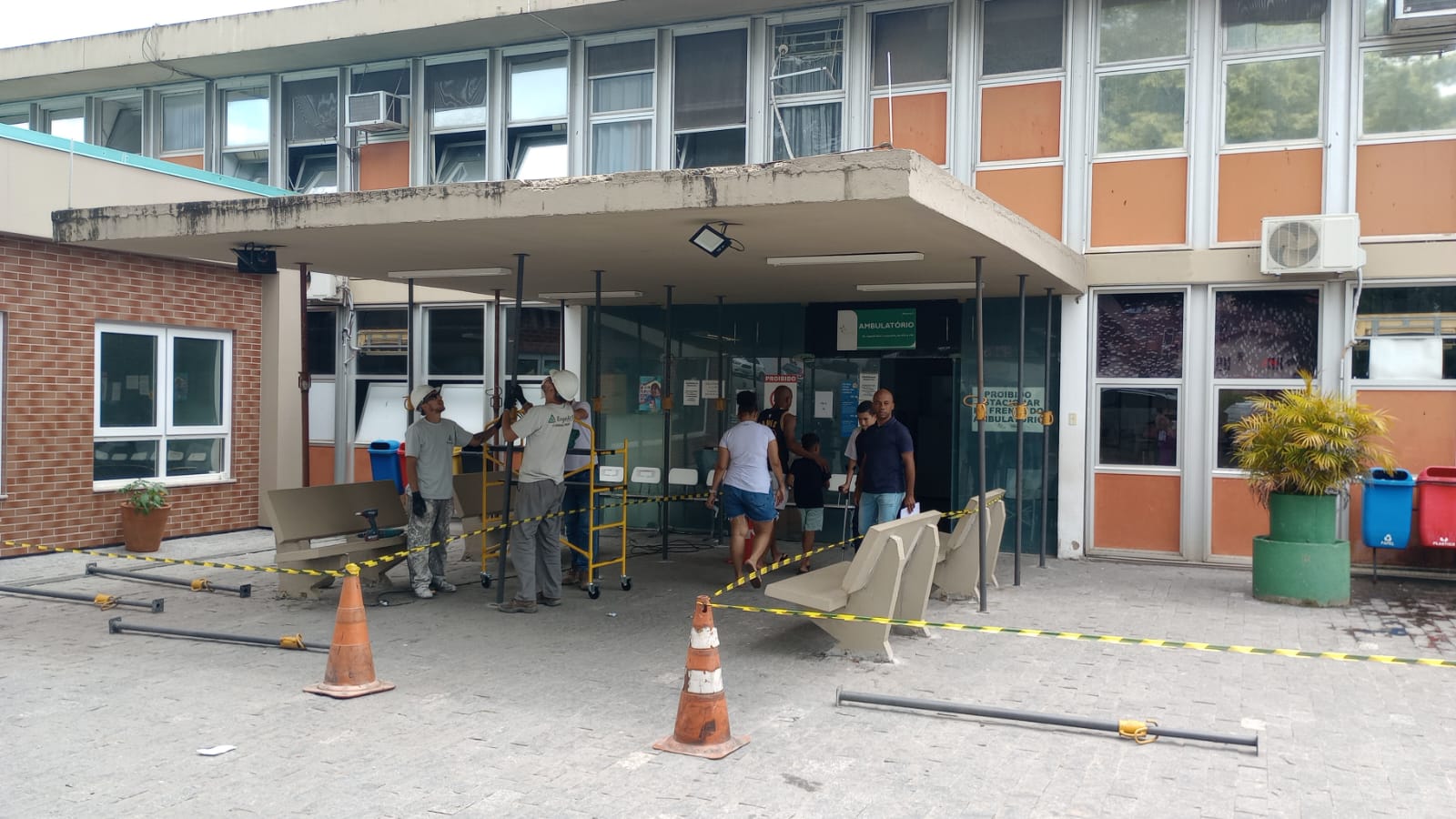 A imagem mostra a entrada de um prédio hospitalar, identificada como setor de ambulatório, passando por manutenção na marquise. Dois trabalhadores, equipados com capacetes e uniforme, realizam o serviço usando andaimes e ferramentas, enquanto a área está isolada por fitas de segurança e cones laranja. Algumas pessoas, incluindo adultos e crianças, entram ou saem pela porta de vidro, e cadeiras de espera foram deslocadas para liberar o espaço de trabalho. O ambiente externo apresenta paredes de tijolos e janelas amplas, com lixeiras coloridas e vasos de plantas decorando o local.