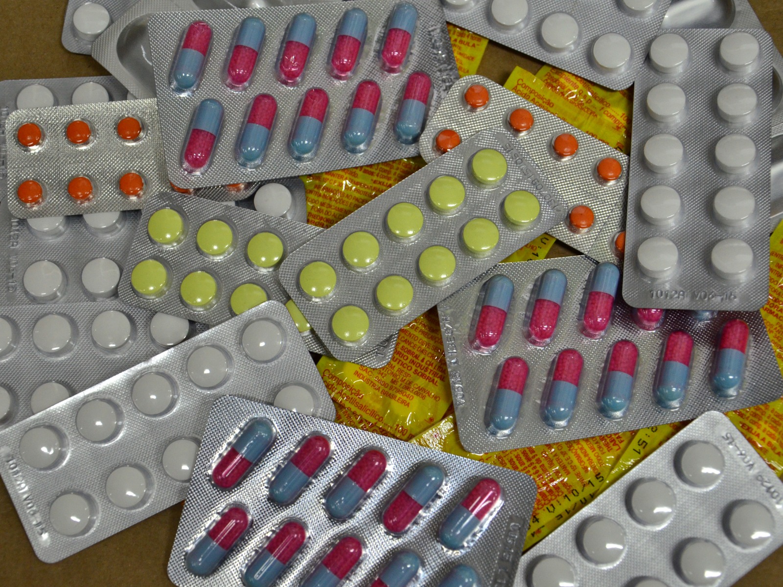 Diversas cartelas de comprimidos e cápsulas em diferentes cores e formatos, como brancas, amarelas, alaranjadas e cápsulas rosa com azul, espalhadas sobre uma superfície com embalagens amarelas de fundo. Os medicamentos estão em blisters prateados.