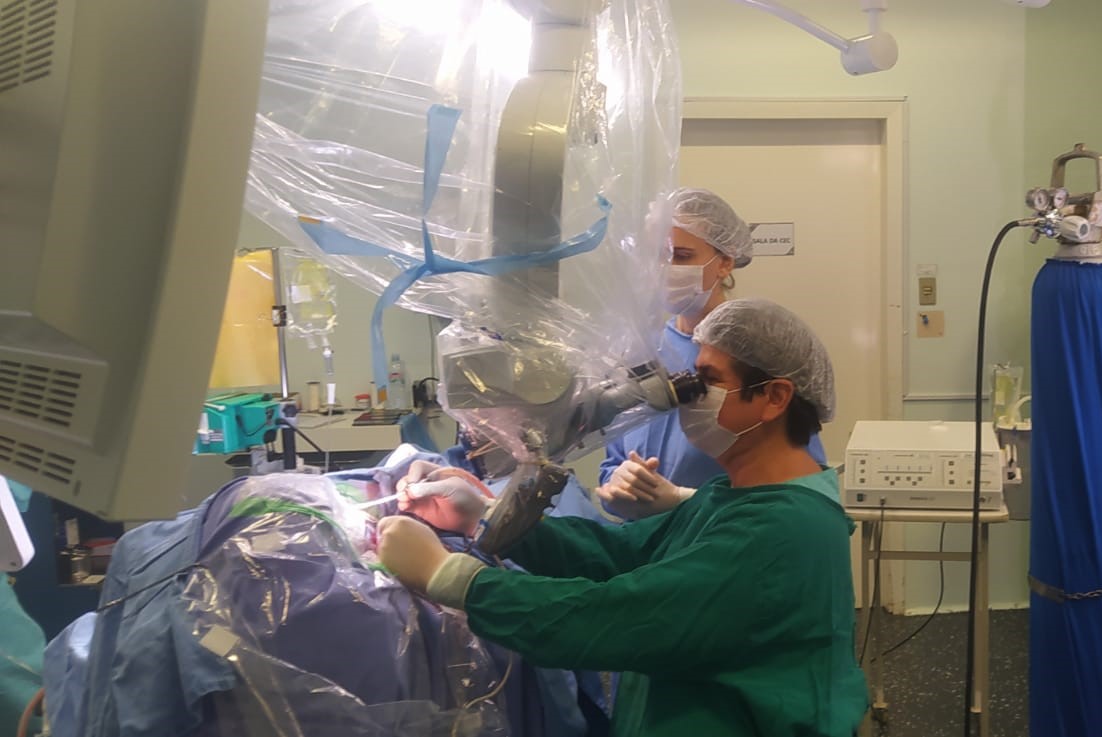 A imagem mostra uma equipe médica realizando um procedimento de neurocirurgia em um centro cirúrgico. Podemos observar que o cirurgião principal utiliza um microscópio cirúrgico, equipamento essencial para realizar intervenções de alta precisão no cérebro ou na medula espinhal. O campo cirúrgico está protegido por campos estéreis e coberto com plástico transparente para manter a assepsia do equipamento.