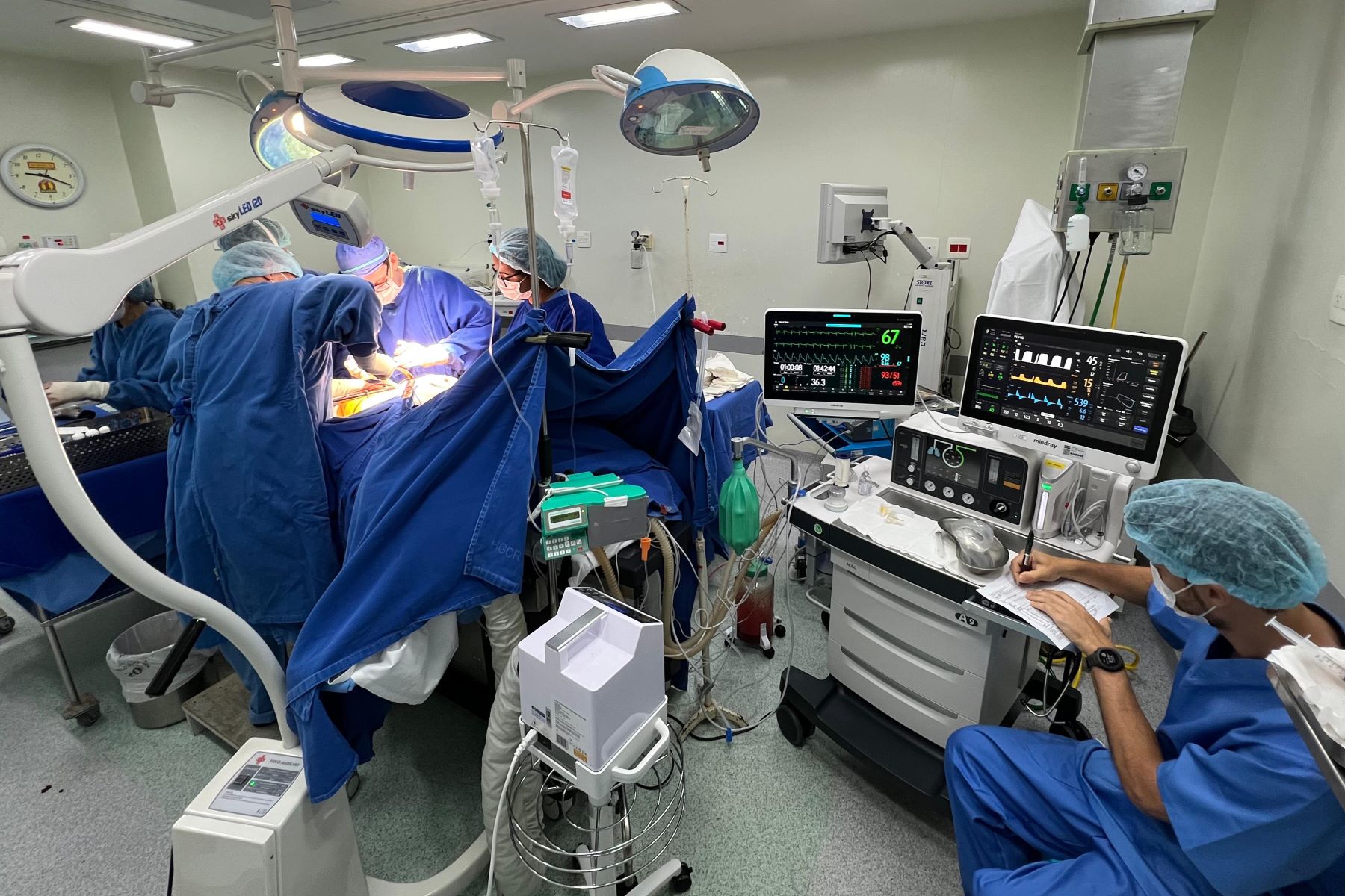 A imagem mostra uma equipe médica realizando um procedimento cirúrgico em um centro cirúrgico totalmente equipado.  No lado esquerdo, vários profissionais — usando aventais, toucas e máscaras cirúrgicas azuis — estão concentrados na cirurgia, com iluminação intensa vinda de refletores cirúrgicos suspensos. Há também um equipamento de imagem médica (braço articulado) posicionado próximo ao paciente.  No lado direito, um profissional está sentado monitorando e registrando informações, com dois monitores exibindo sinais vitais como frequência cardíaca, pressão arterial, saturação de oxigênio e outros parâmetros. Ao redor, há bombas de infusão, respiradores, cilindros e equipamentos de suporte à vida, todos conectados ao paciente.  O ambiente é típico de um hospital, com piso lavável, paredes claras e organização pensada para manter a assepsia e segurança do procedimento.