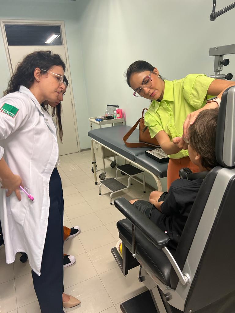 Em uma sala de atendimento oftalmológico, uma profissional de camisa verde e óculos transparentes examina o olho de uma criança sentada em uma cadeira de atendimento médico. Ao lado, outra profissional de jaleco branco com logotipo no braço esquerdo observa atentamente o procedimento. Ao fundo, há uma maca, um banco com degraus e equipamentos médicos.