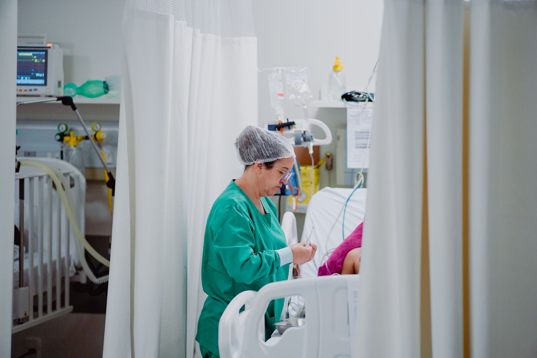 Profissional de saúde com touca e jaleco verde preparando materiais ao lado de paciente deitado em leito hospitalar, durante mutirão de cateterismo. O ambiente é delimitado por cortinas brancas, com equipamentos médicos ao fundo.