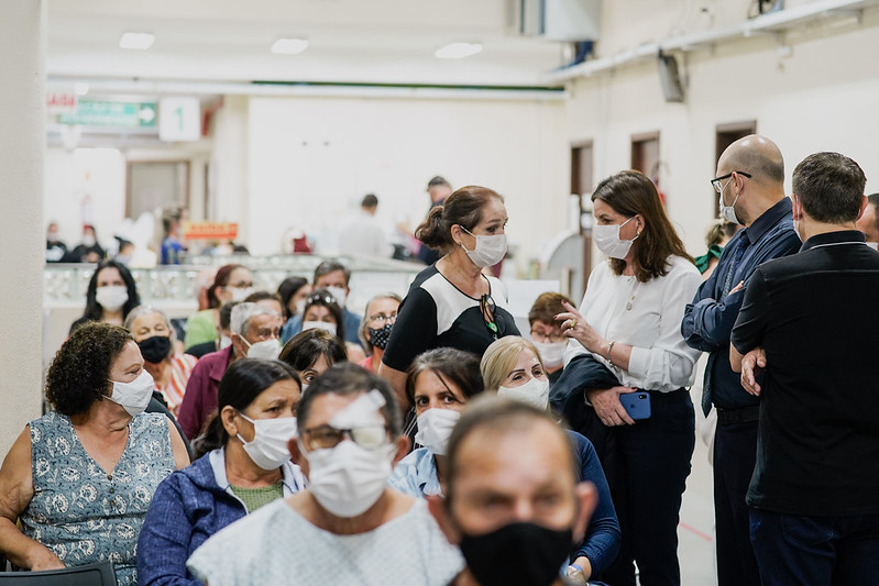 Sala de espera de hospital com dezenas de pessoas, a maioria usando máscaras. Alguns pacientes têm curativos nos olhos, indicando recuperação de cirurgias oftalmológicas, como catarata. Profissionais de saúde conversam com o público, enquanto outros circulam ao fundo. Cena representa atendimento coletivo em um mutirão de saúde.