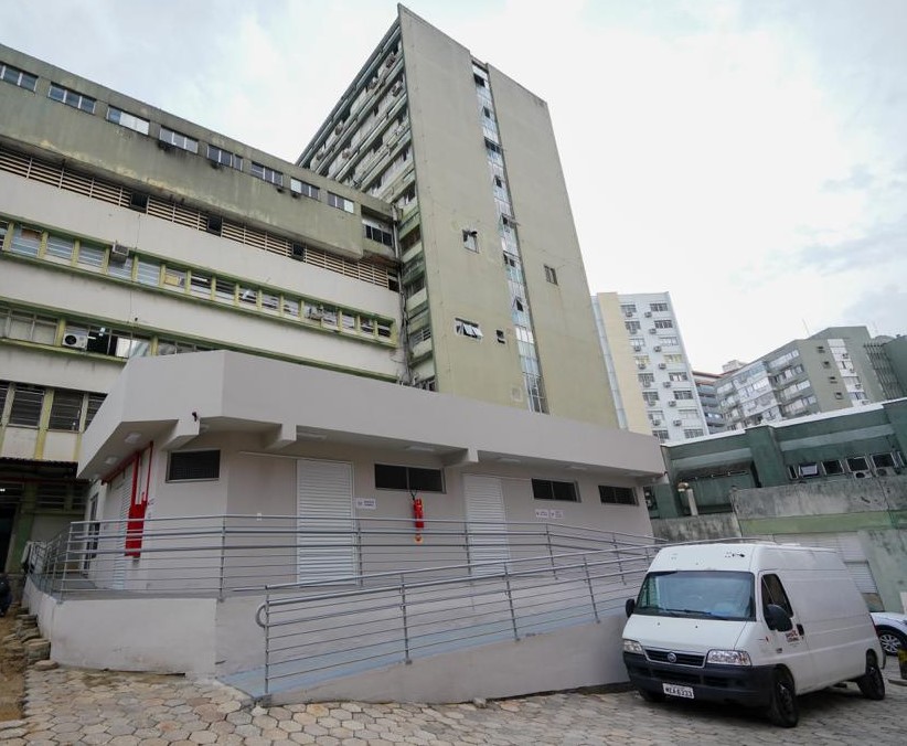 Vista externa do novo abrigo de resíduos do Hospital Governador Celso Ramos, com estrutura térrea recém-construída, rampa de acesso com corrimão metálico e portas metálicas fechadas. Um furgão branco está estacionado em frente ao prédio.