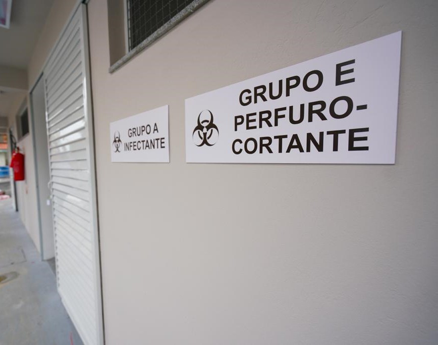 Vista de uma parede externa com sinalização para descarte de resíduos hospitalares, com placas indicando "Grupo A Infectante" e "Grupo E Perfurocortante", ambas com o símbolo de risco biológico. À esquerda, portas metálicas e extintores de incêndio são visíveis ao longo do corredor.
