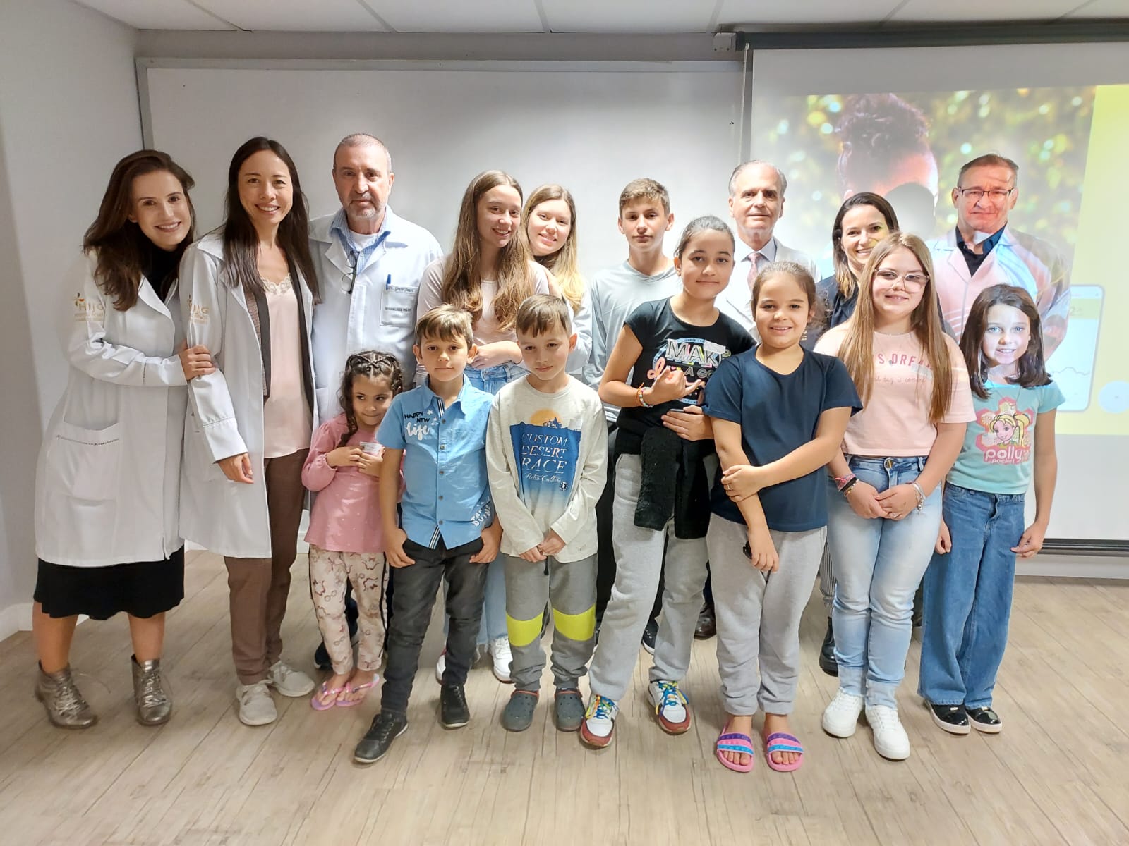 Grupo formado por médicos, crianças e adolescentes posa para foto em sala de aula ou auditório. Três profissionais de saúde com jaleco branco estão à esquerda, acompanhados por cerca de 14 crianças e adolescentes sorridentes. Ao fundo, uma tela de projeção exibe a imagem de um homem idoso com jaleco esportivo, ao lado de gráficos de um aplicativo de monitoramento de saúde, possivelmente relacionado a sensor de glicose.