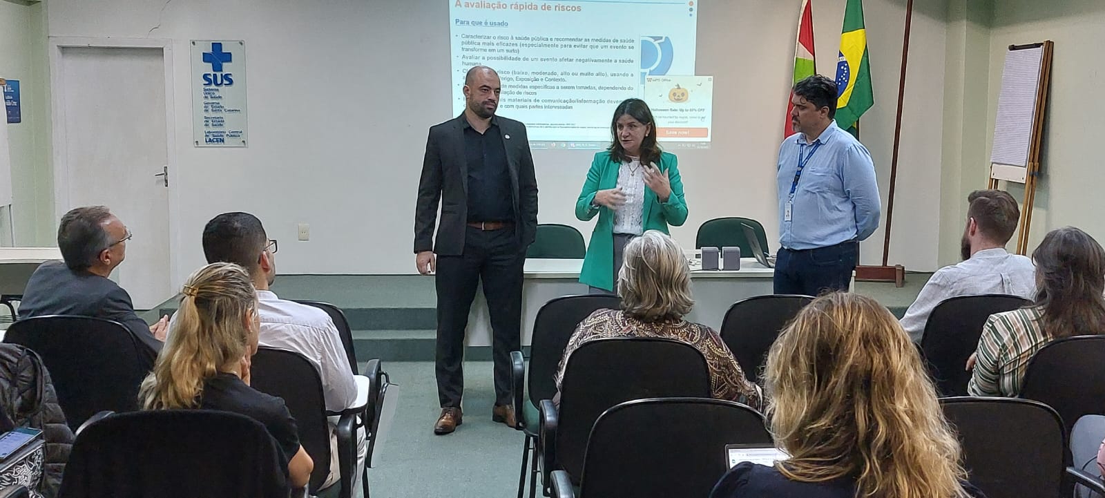 Três palestrantes em frente a uma tela de projeção durante oficina sobre avaliação rápida de riscos à saúde pública, com participantes sentados e materiais do SUS visíveis na sala.