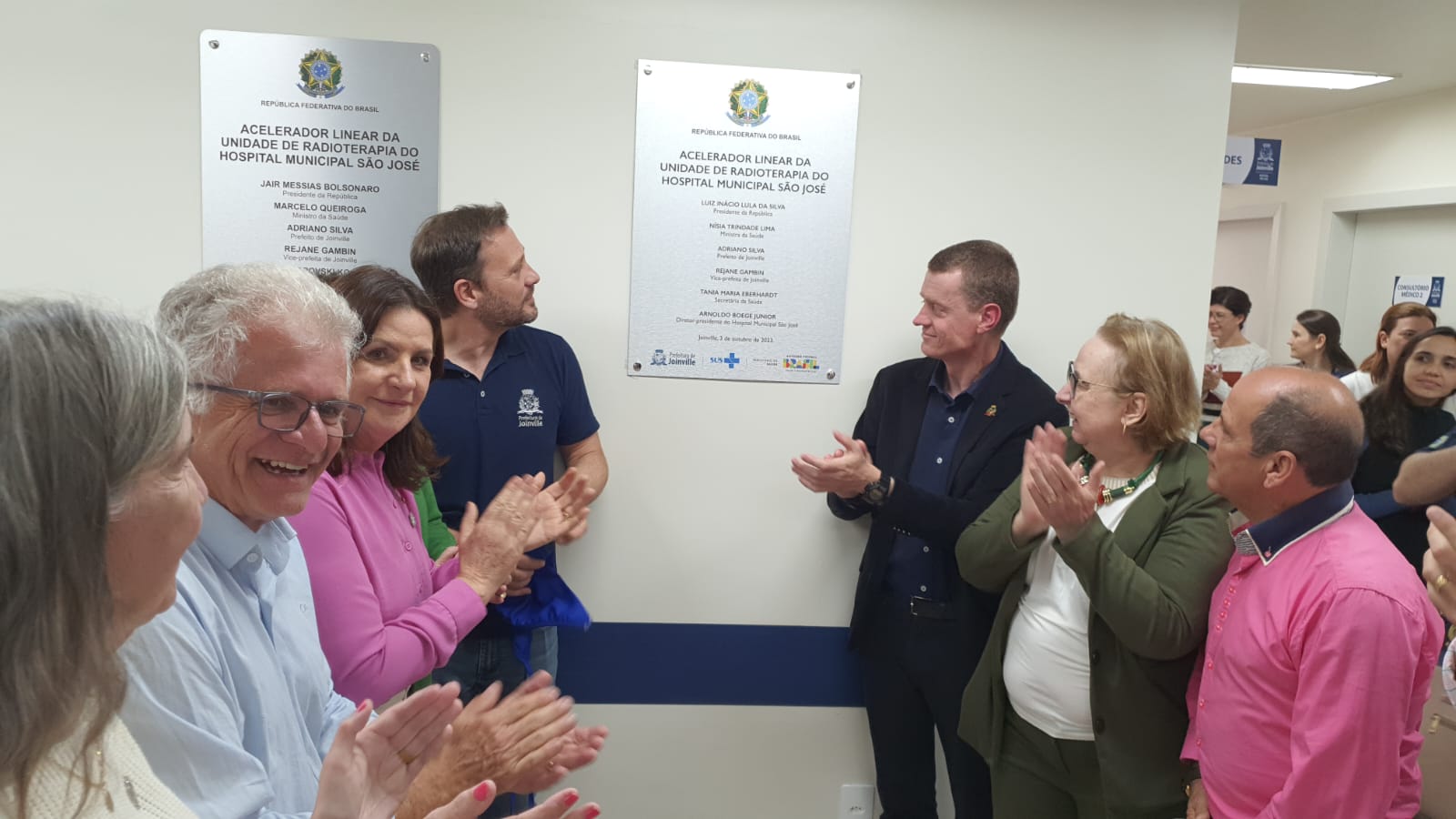 Grupo de pessoas aplaude em frente a duas placas inaugurais fixadas na parede, marcando a entrega do acelerador linear da Unidade de Radioterapia do Hospital Municipal São José. Participam autoridades políticas e administrativas, todos demonstrando satisfação durante a cerimônia.