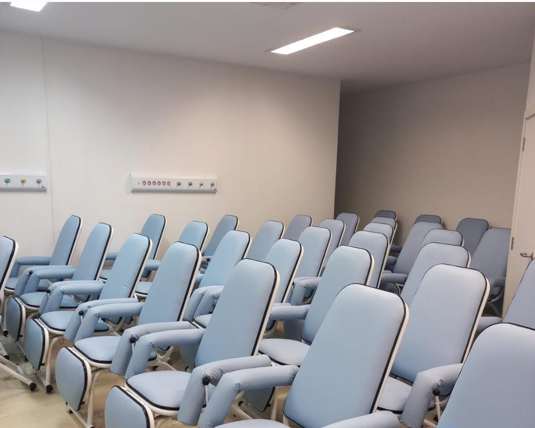 Sala equipada com várias poltronas hospitalares azuis dispostas em fileiras, voltadas para a mesma direção, em ambiente iluminado e climatizado.