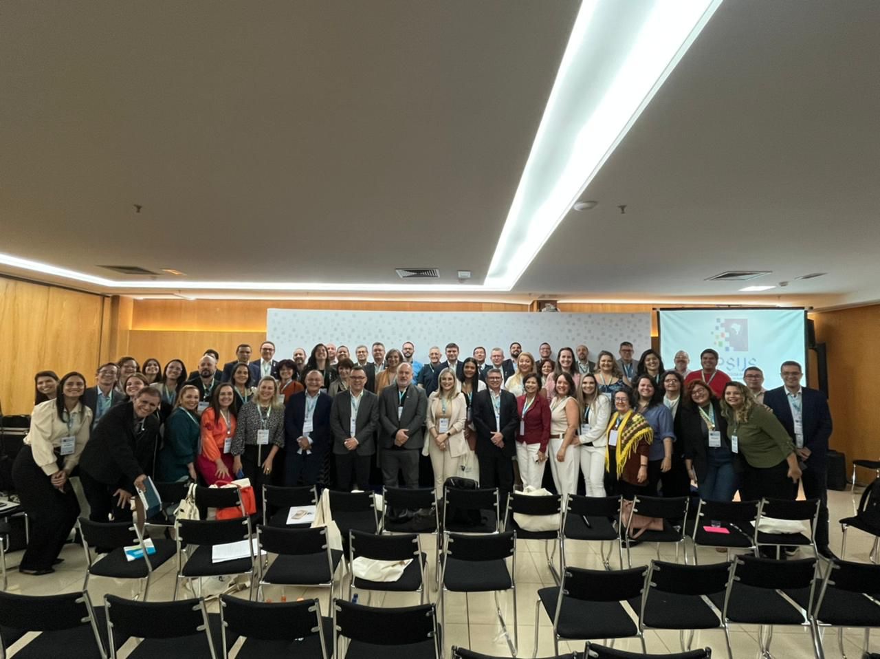 Imagem de um grande grupo de pessoas posando para foto em ambiente fechado, possivelmente uma conferência ou encontro institucional. Ao fundo há um painel com a logomarca do PPSUS projetada em uma tela. Os participantes usam crachás e trajes formais ou sociais.