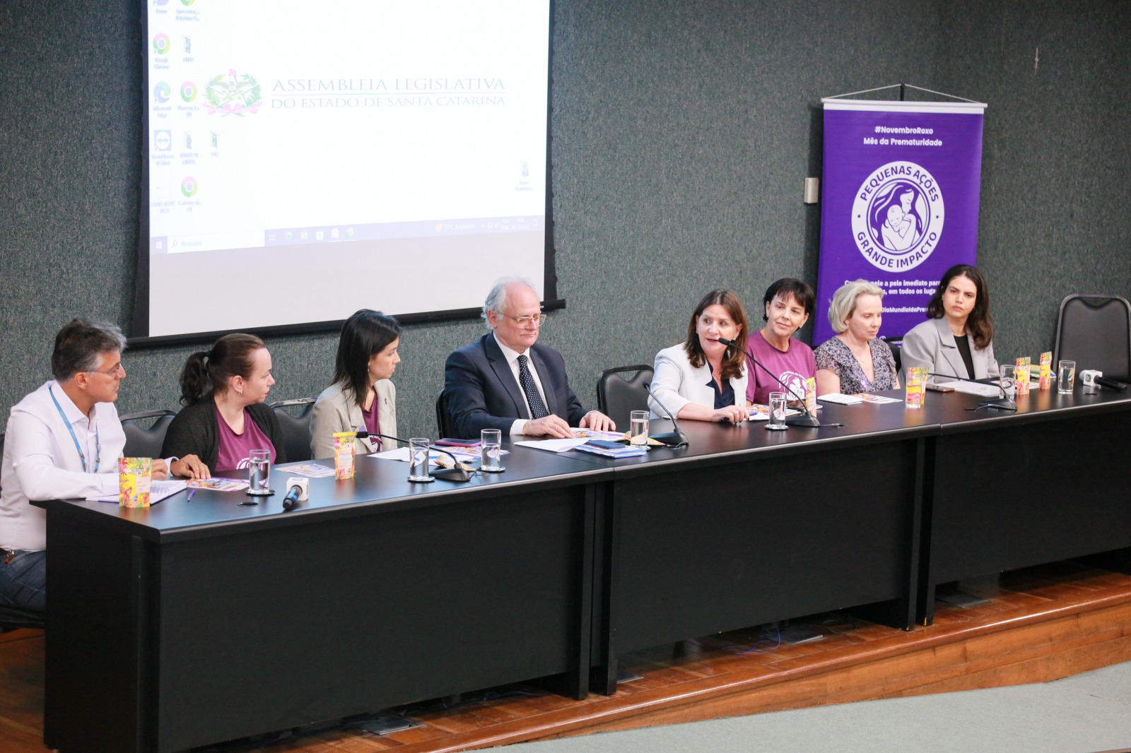 A imagem mostra uma mesa de conferência com oito pessoas sentadas lado a lado, participando de um evento na Assembleia Legislativa do Estado de Santa Catarina.  Primeiro plano: Todos estão voltados para o público, com microfones, copos de água, sucos e folhetos à frente.  Centro: Entre os participantes, há homens e mulheres, alguns vestidos formalmente, outros com camisetas roxas alusivas à causa do evento. Uma das mulheres fala ao microfone, parecendo conduzir a pauta.  Fundo: À esquerda, o telão exibe o brasão e o nome da Assembleia Legislativa. À direita, um banner roxo destaca a campanha “Novembro Roxo – Mês da Prematuridade”, com o lema “Pequenas ações, grande impacto” e ilustração de um bebê nos braços.