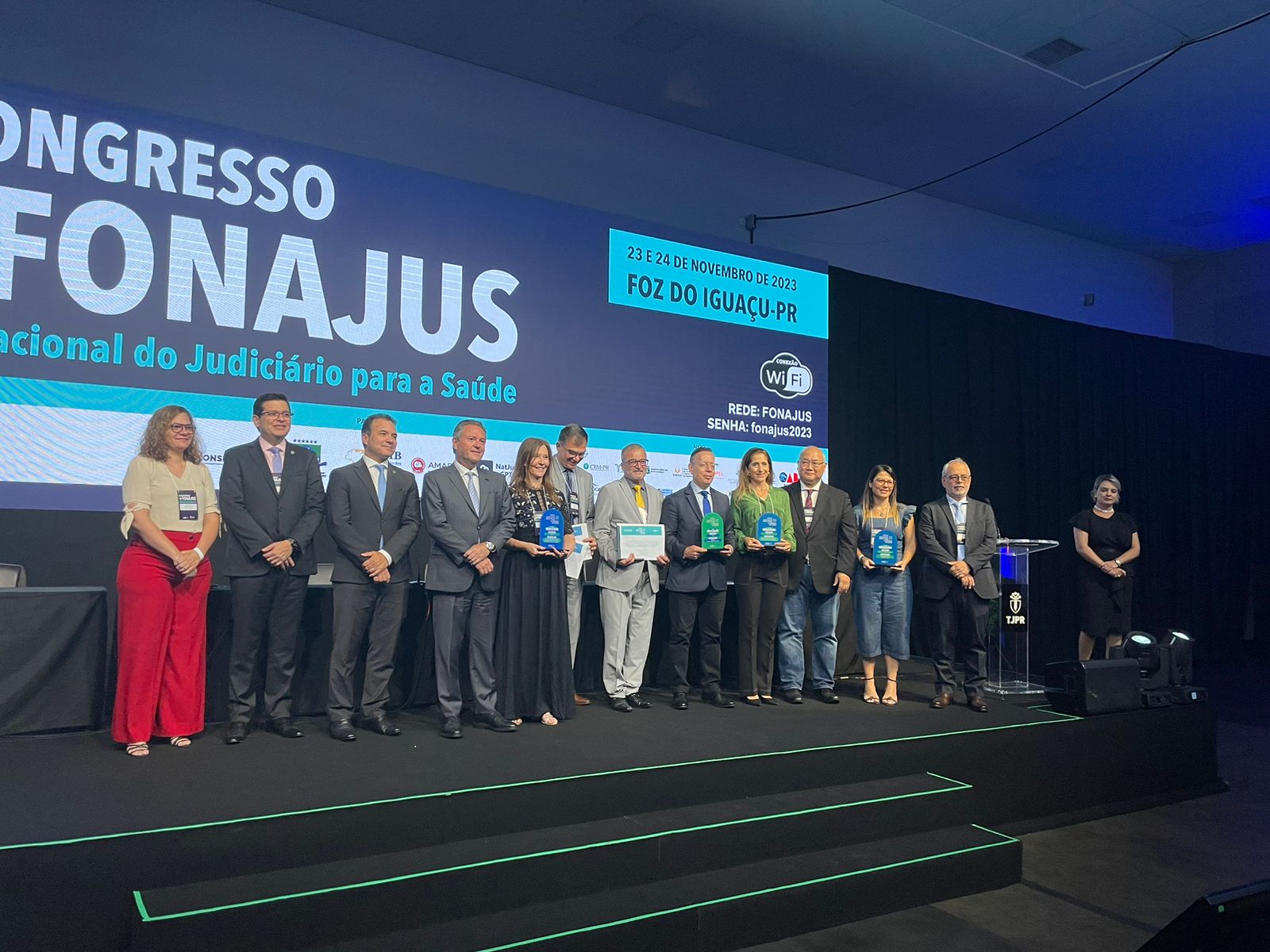 Grupo de pessoas em um palco durante a premiação no Congresso Nacional do Judiciário para a Saúde (FONAJUS), realizado em Foz do Iguaçu-PR nos dias 23 e 24 de novembro de 2023. Alguns participantes seguram troféus e certificados, enquanto posam para foto diante de um telão com o nome e informações do evento.
