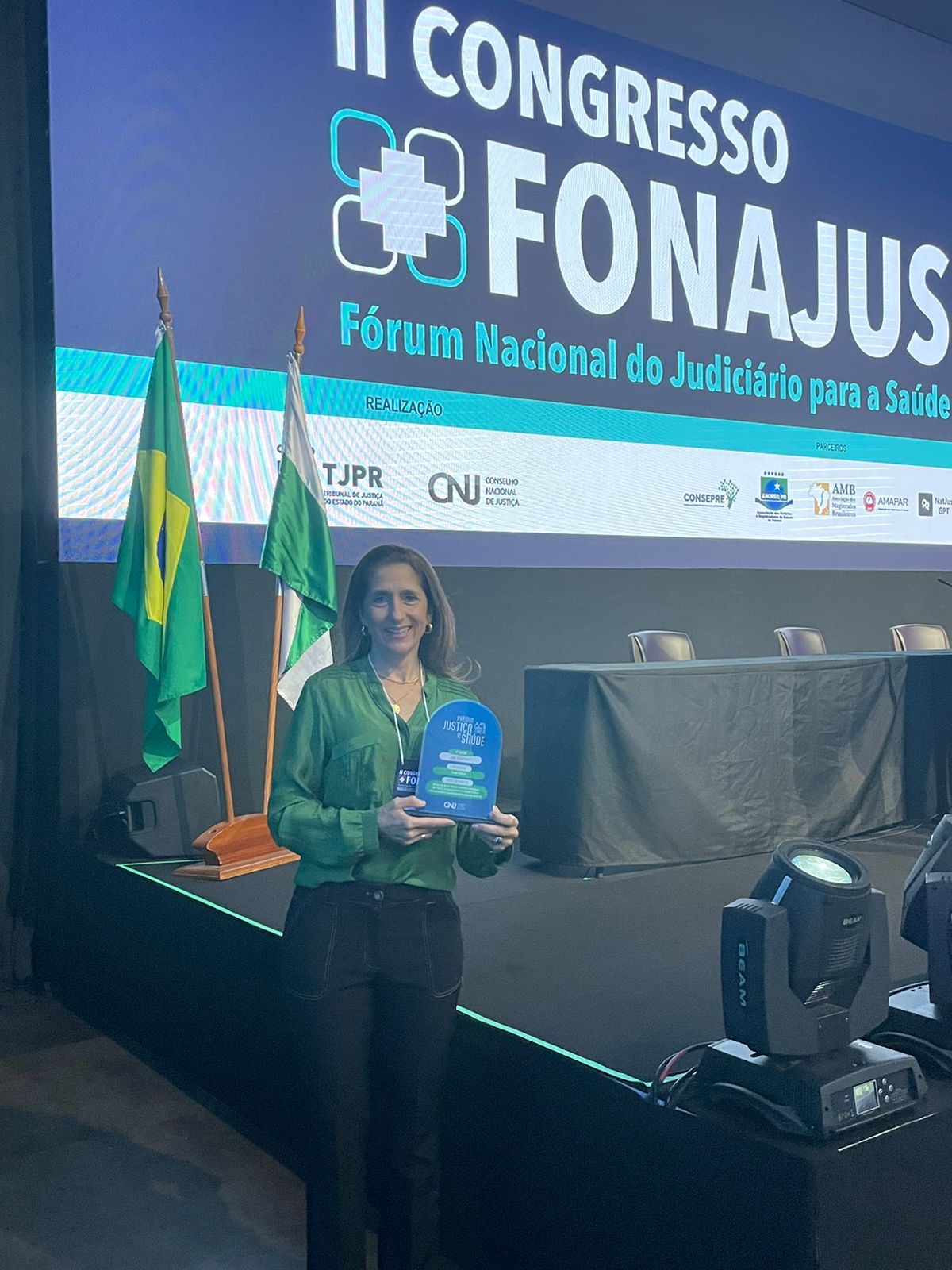 Mulher em pé segurando um troféu azul do Prêmio Fórum Nacional do Judiciário para a Saúde, durante o II Congresso FONAJUS. Ao fundo, painel com o nome do evento, bandeiras do Brasil e do Paraná, e parte do palco com cadeiras e iluminação de evento.