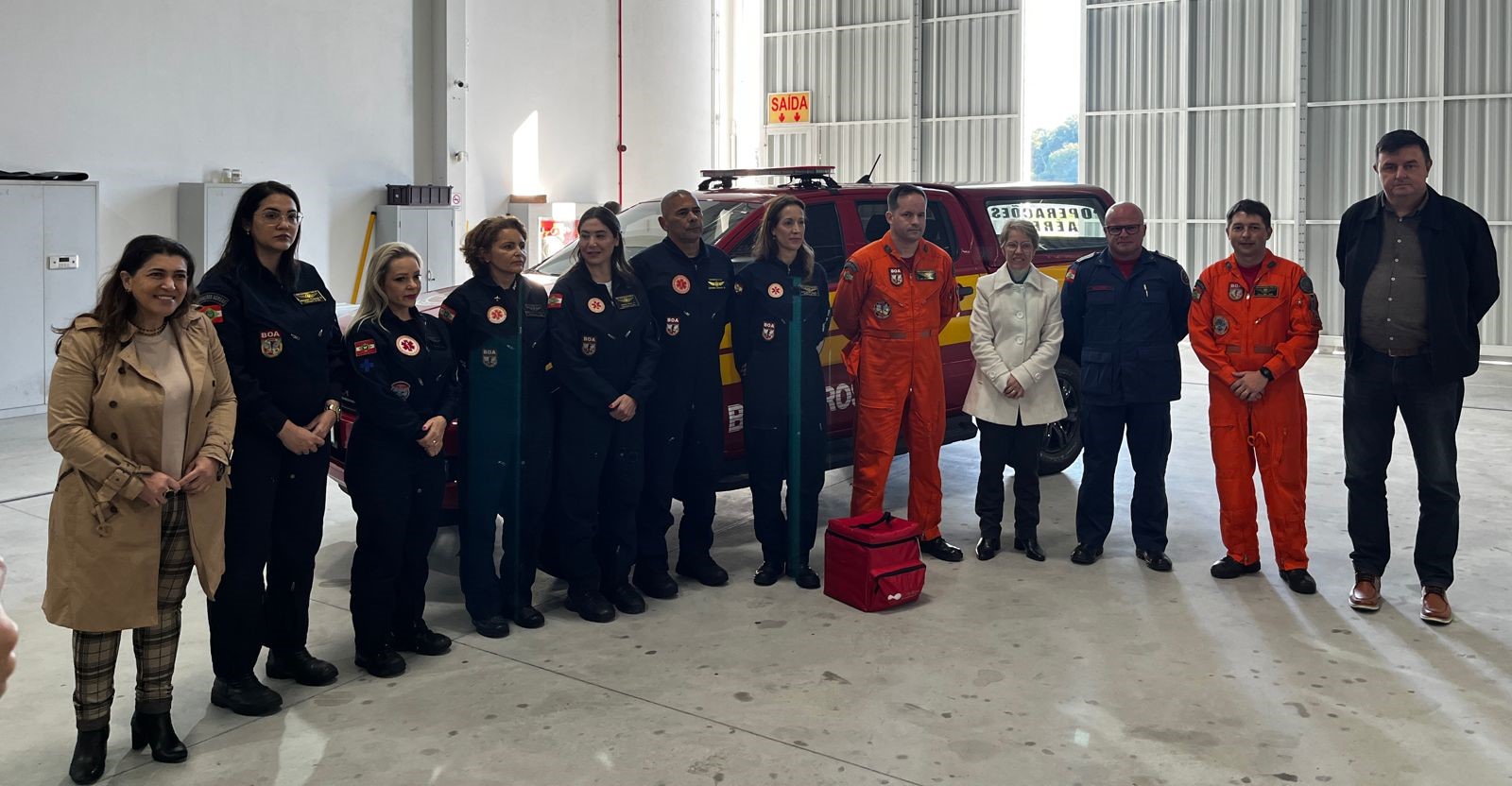 Grupo de profissionais da saúde e do Corpo de Bombeiros posando para foto em hangar, com viatura de resgate ao fundo. Alguns vestem uniformes pretos com brasões do SAMU e BOA, outros estão em macacões laranja e roupas civis. Evento relacionado ao programa Sangue Total.