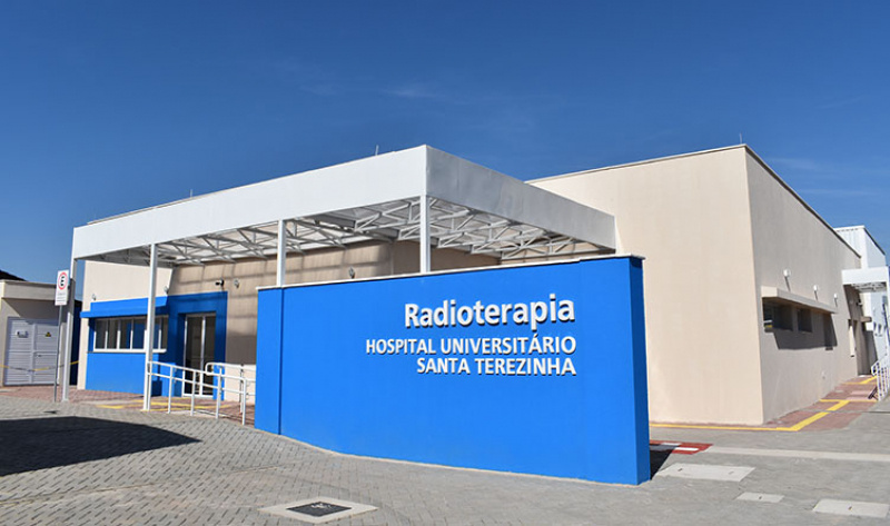 Fachada do setor de Radioterapia do Hospital Universitário Santa Terezinha (HUST), com paredes em tons claros e um letreiro azul em destaque com o nome do serviço. O prédio é moderno, com acesso por rampa e cobertura metálica branca. O céu está limpo, evidenciando um dia ensolarado.