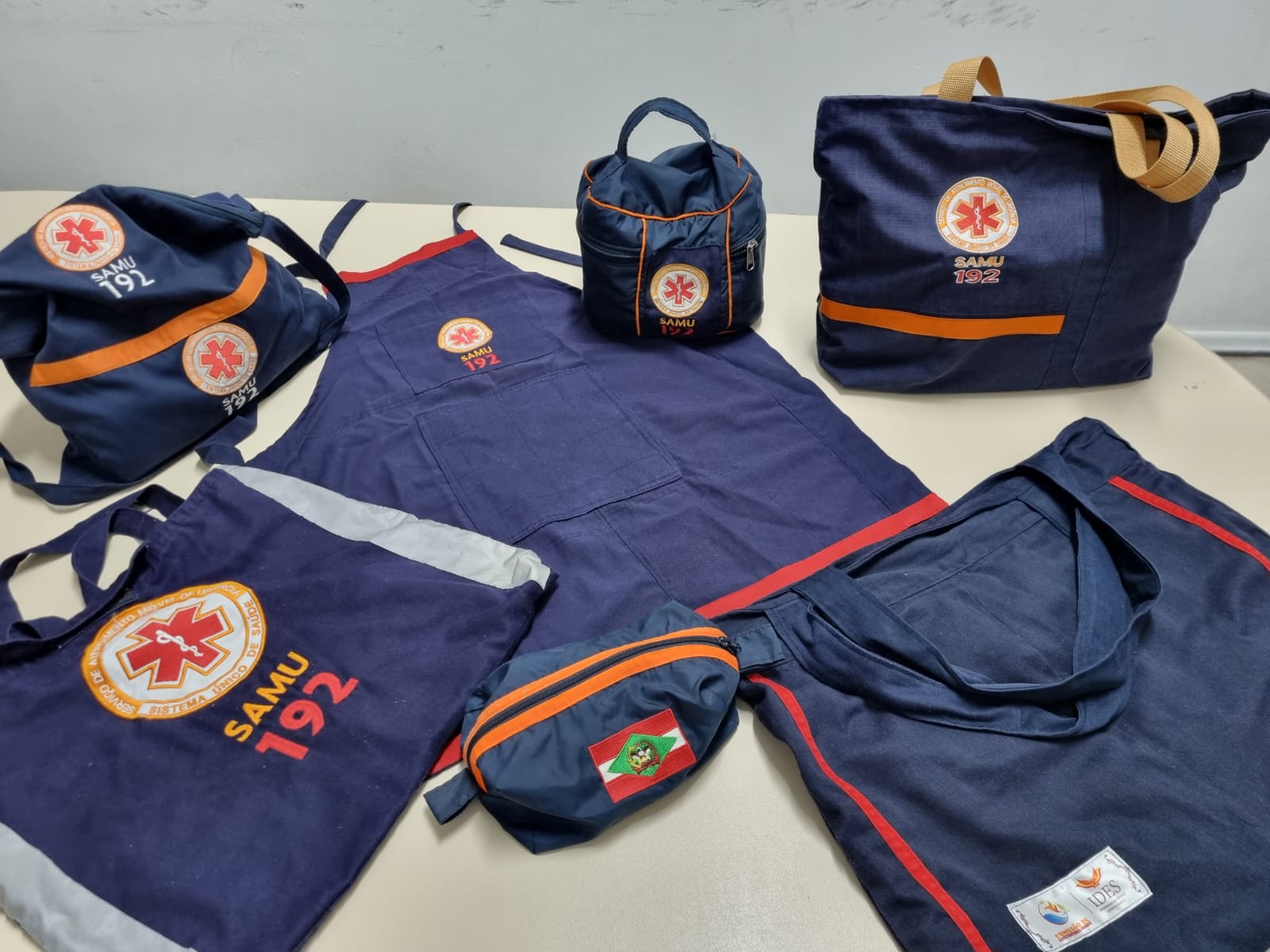 Diversos itens confeccionados em tecido azul marinho com detalhes em laranja, vermelho e cinza estão dispostos sobre uma mesa clara. Os itens incluem bolsas, aventais e nécessaires com o logotipo do SAMU 192 (Serviço de Atendimento Móvel de Urgência) e do SUS. Um dos objetos também apresenta a bandeira de Santa Catarina, e outro exibe a logomarca do IDES (Instituto de Desenvolvimento, Ensino e Assistência à Saúde). Todos os itens parecem ser produtos reciclados ou reaproveitados a partir de uniformes e materiais institucionais.