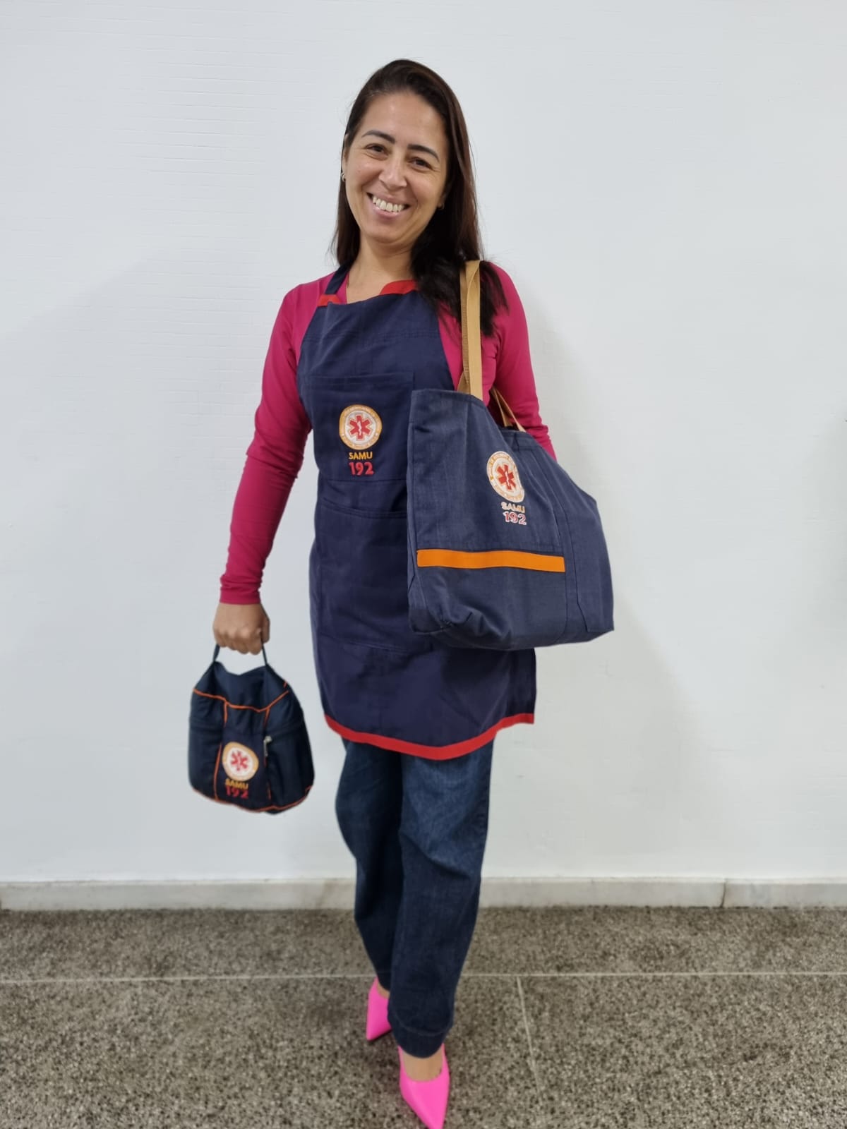 Mulher sorridente posa de frente para a câmera, vestindo blusa rosa de manga longa, jeans e sapatos de salto alto rosa vibrante. Ela usa um avental azul marinho com detalhes vermelhos e o logotipo do SAMU 192 bordado no centro. Carrega uma bolsa grande no ombro esquerdo e uma nécessaire pequena na mão direita, ambos feitos do mesmo tecido do avental e com o mesmo logotipo. O fundo é uma parede branca e o piso é de granito escuro.