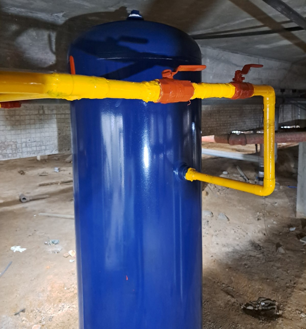 Cilindro azul metálico com tubulação amarela conectada, com registros de controle na parte superior. Equipamento instalado em ambiente subterrâneo com piso de terra batida e teto de concreto.