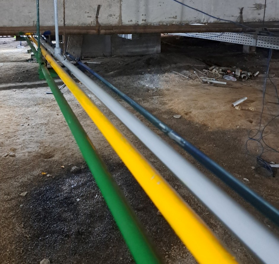 Tubulações de gases medicinais instaladas em ambiente subterrâneo, fixadas em suporte metálico. Os tubos são pintados em verde, amarelo, cinza e preto, indicando diferentes tipos de gases.