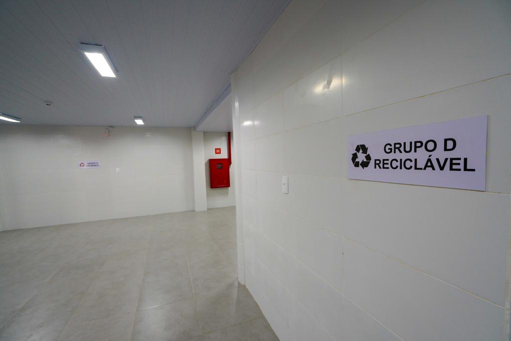 Ambiente interno com paredes revestidas de azulejos brancos e piso em porcelanato, com placas indicativas de descarte de resíduos, incluindo "Grupo D Reciclável". No fundo, há um hidrante vermelho instalado na parede.