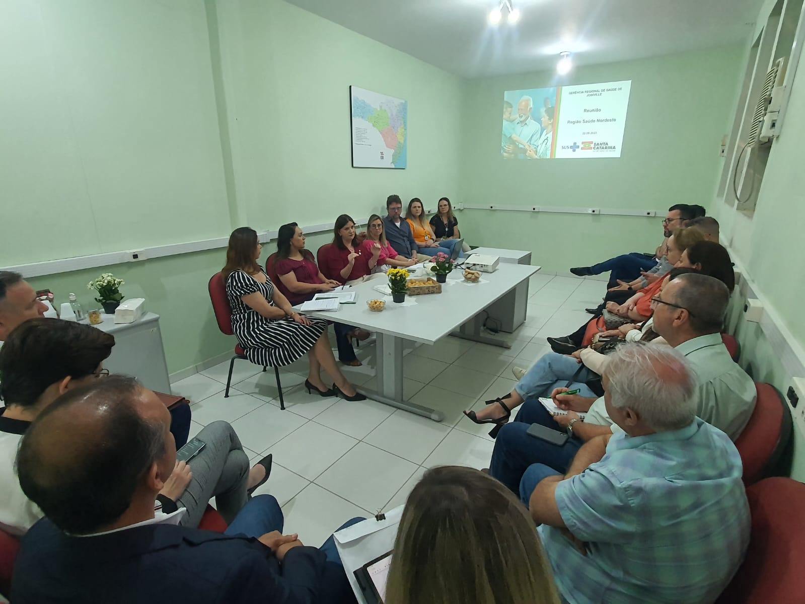 Reunião em uma sala com paredes verdes claras e piso branco, onde cerca de vinte pessoas estão sentadas em cadeiras ao redor de uma mesa retangular. Uma mulher ao centro fala, enquanto os demais escutam atentamente. Na parede ao fundo, uma projeção exibe o título "Reunião Região Sul e Nordeste" com os logotipos da SES e Governo de Santa Catarina. Há flores, café e petiscos sobre a mesa.