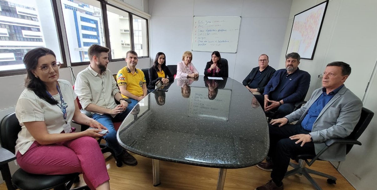 A imagem mostra uma reunião com nove pessoas sentadas ao redor de uma mesa de reuniões preta e retangular, em uma sala com janelas amplas que permitem a entrada de luz natural. À esquerda, há quatro pessoas, incluindo uma mulher de óculos e calça rosa na ponta da mesa, seguida por um homem de camisa branca, um homem com camisa polo amarela e uma mulher de blusa preta. No centro e à direita, há cinco pessoas, incluindo uma mulher de blusa rosa, outra mulher de blazer preto, e três homens de camisa social, sendo um deles de blazer cinza. Ao fundo, há um quadro branco com anotações e um quadro colorido na parede.