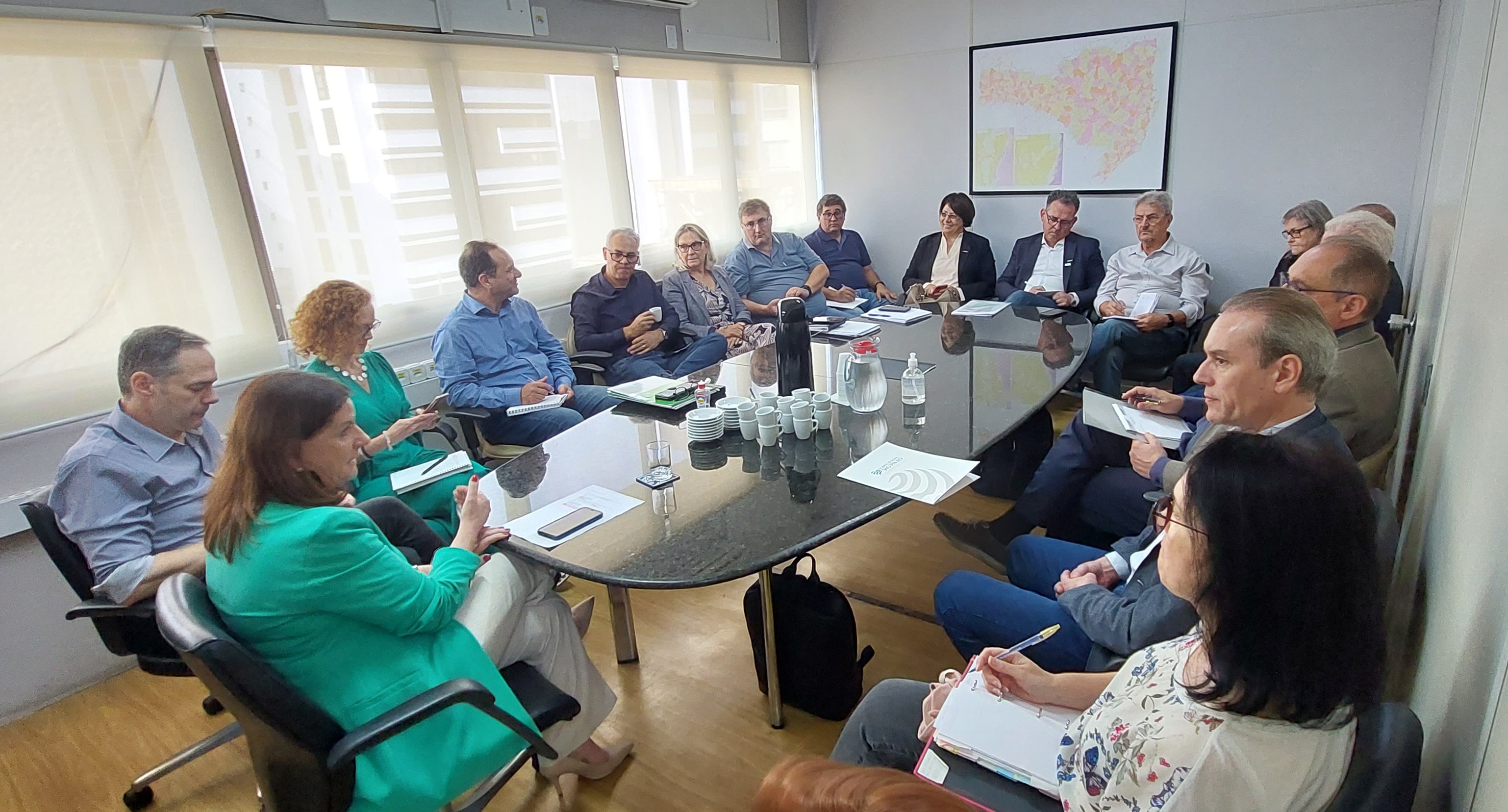 Grupo de aproximadamente vinte pessoas reunido em torno de uma mesa oval em uma sala de reuniões iluminada por janelas. Os participantes, homens e mulheres, estão sentados com cadernos, papéis e copos à mesa, em ambiente formal. Ao fundo, há um mapa colorido na parede e materiais de escritório.