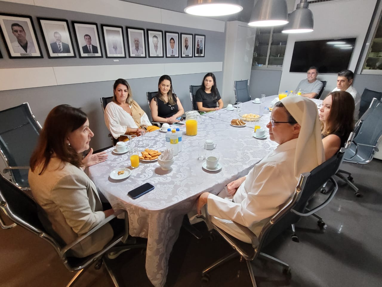 Reunião formal em uma sala com cerca de dez pessoas sentadas ao redor de uma mesa coberta com toalha branca decorada. A mesa contém sucos, biscoitos e xícaras de café. Uma religiosa com hábito branco está entre os participantes, ao lado de uma mulher que parece conduzir a conversa. Ao fundo, há quadros com retratos de profissionais pendurados na parede.