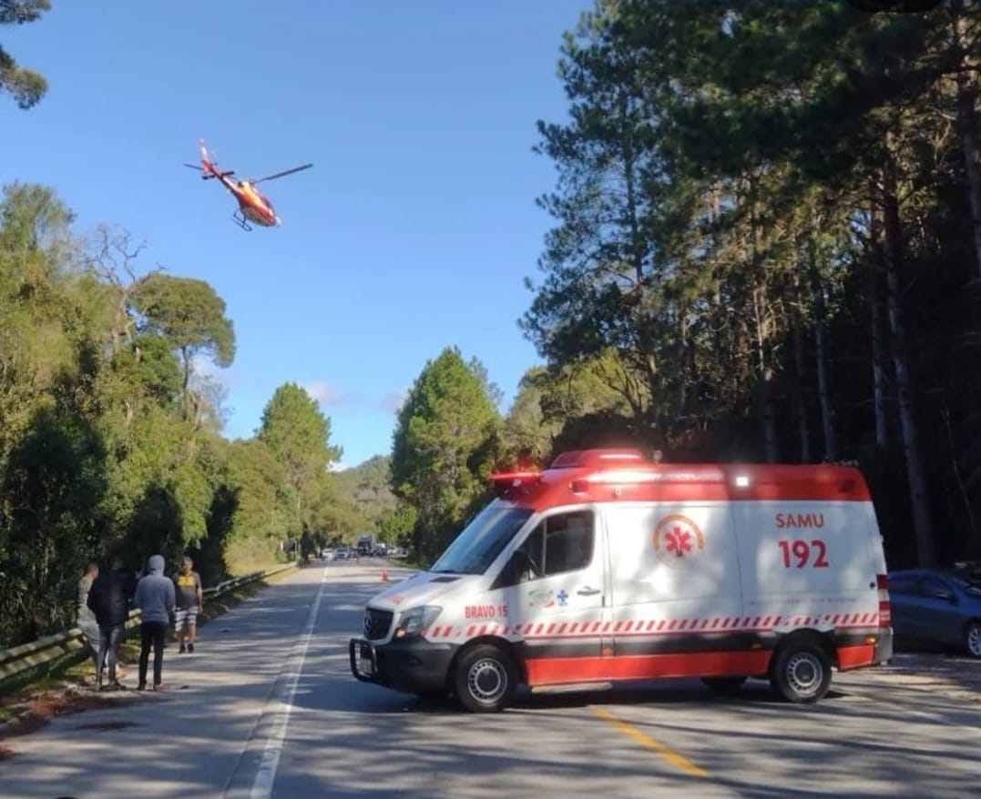 A imagem mostra uma cena de atendimento de emergência em uma rodovia cercada por árvores.  Em primeiro plano, há uma ambulância do SAMU 192, modelo van, estacionada atravessada na pista para bloquear o tráfego.  Ao fundo, um helicóptero de resgate sobrevoa a área, provavelmente se aproximando para pousar.  Três pessoas estão paradas no acostamento, observando a movimentação.  O céu está limpo e ensolarado, indicando que a ocorrência acontece em condições climáticas favoráveis.  O cenário sugere uma operação conjunta de resgate terrestre e aéreo, possivelmente devido a um acidente ou situação médica grave na estrada.