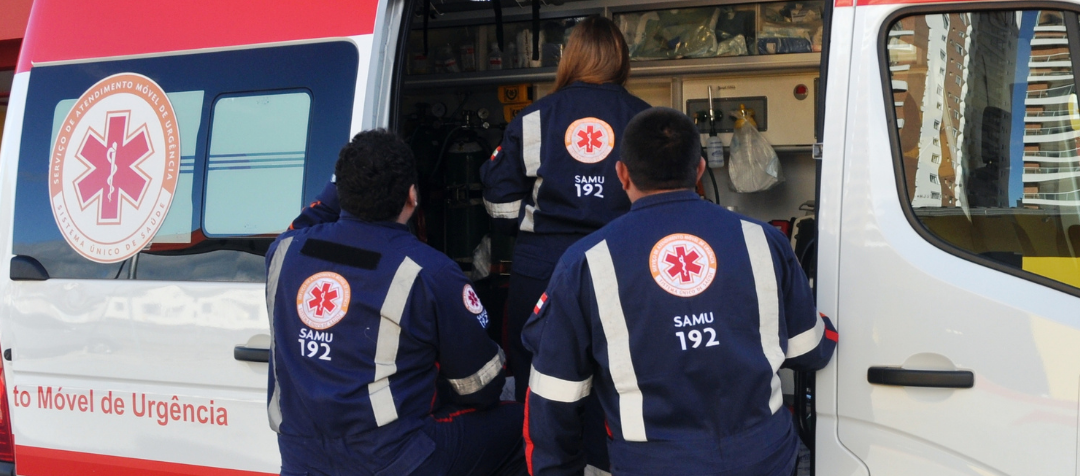 Três profissionais do SAMU 192, usando uniformes azul-marinho com faixas refletivas e o símbolo de emergência nas costas, entrando na parte traseira de uma ambulância equipada para atendimentos de urgência.
