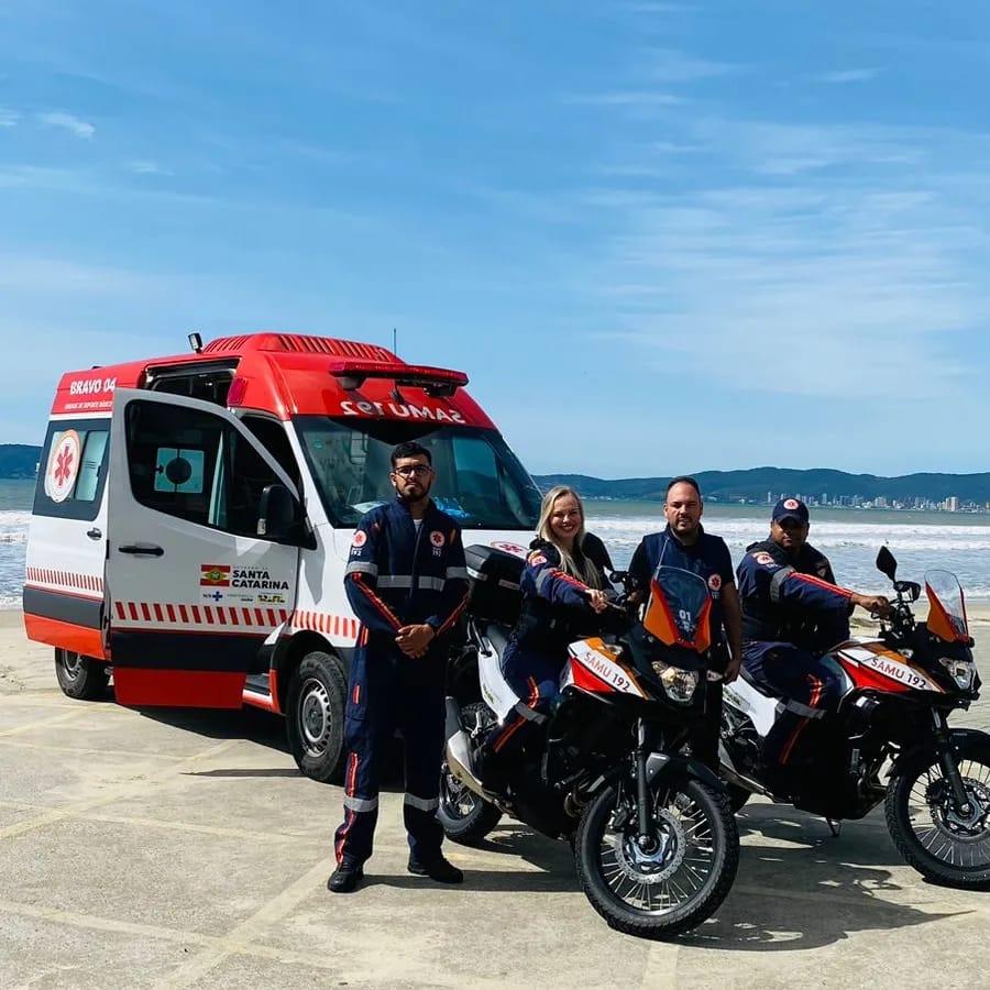 A imagem mostra uma equipe do SAMU 192 posando para foto em um cenário à beira-mar.  Ao fundo, há uma ambulância de suporte básico com a marca do Governo de Santa Catarina, estacionada com a porta lateral aberta.  À frente, quatro profissionais vestindo uniformes azul-marinho com detalhes refletivos e o logotipo do SAMU posam ao lado de duas motocicletas de emergência identificadas com “SAMU 192”.  O grupo está em uma área pavimentada, próxima à praia, com o mar e as montanhas visíveis no horizonte sob um céu parcialmente limpo.  A cena transmite prontidão operacional e integração de diferentes tipos de viaturas para atendimento rápido em áreas urbanas e litorâneas.