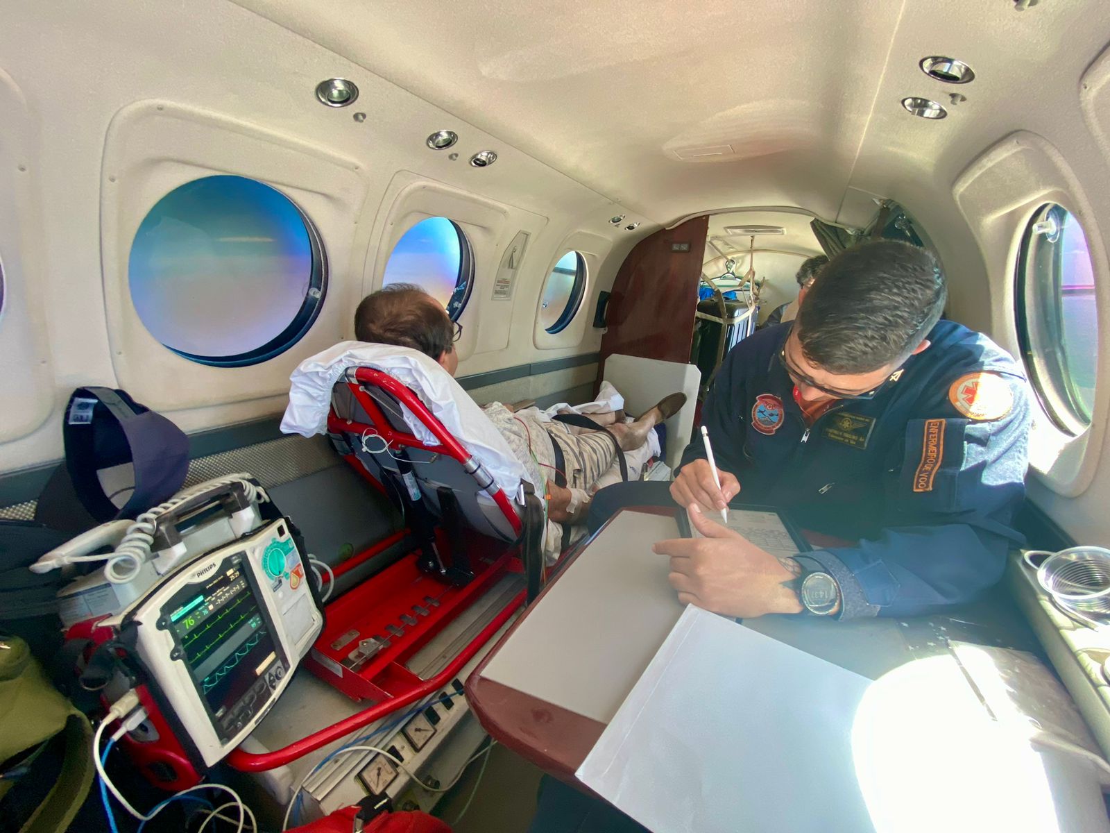 Interior de aeronave de transporte aeromédico, com paciente deitado em maca vermelha próximo à janela, olhando para fora. Ao lado, um monitor cardíaco exibe sinais vitais. Um profissional de saúde, usando uniforme azul com emblemas do SAMU e da aviação, está sentado escrevendo em uma prancheta. A luz do sol entra pelas janelas redondas, iluminando parcialmente a cena.