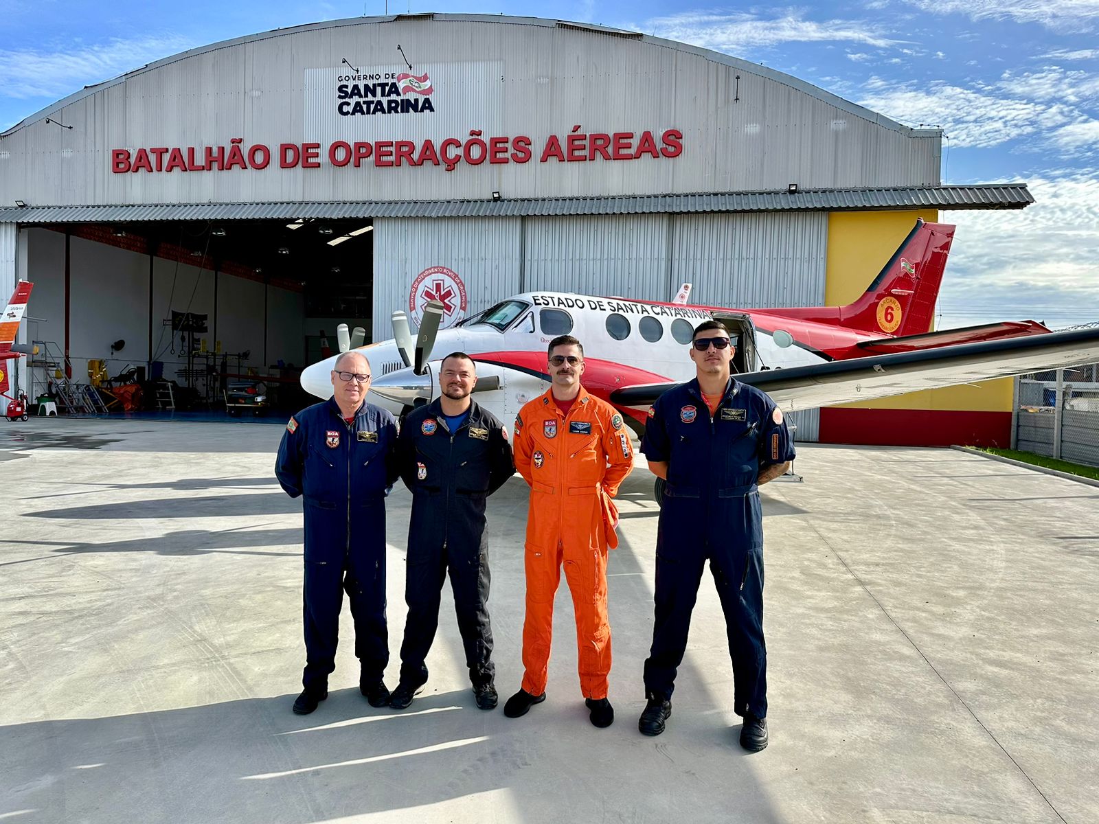 Quatro profissionais do Batalhão de Operações Aéreas de Santa Catarina posam sorridentes em frente a um hangar com a inscrição “Batalhão de Operações Aéreas” e um avião de resgate aeromédico. Três deles vestem macacão azul e um está com macacão laranja. Ao fundo, há outro avião e parte da estrutura do hangar.